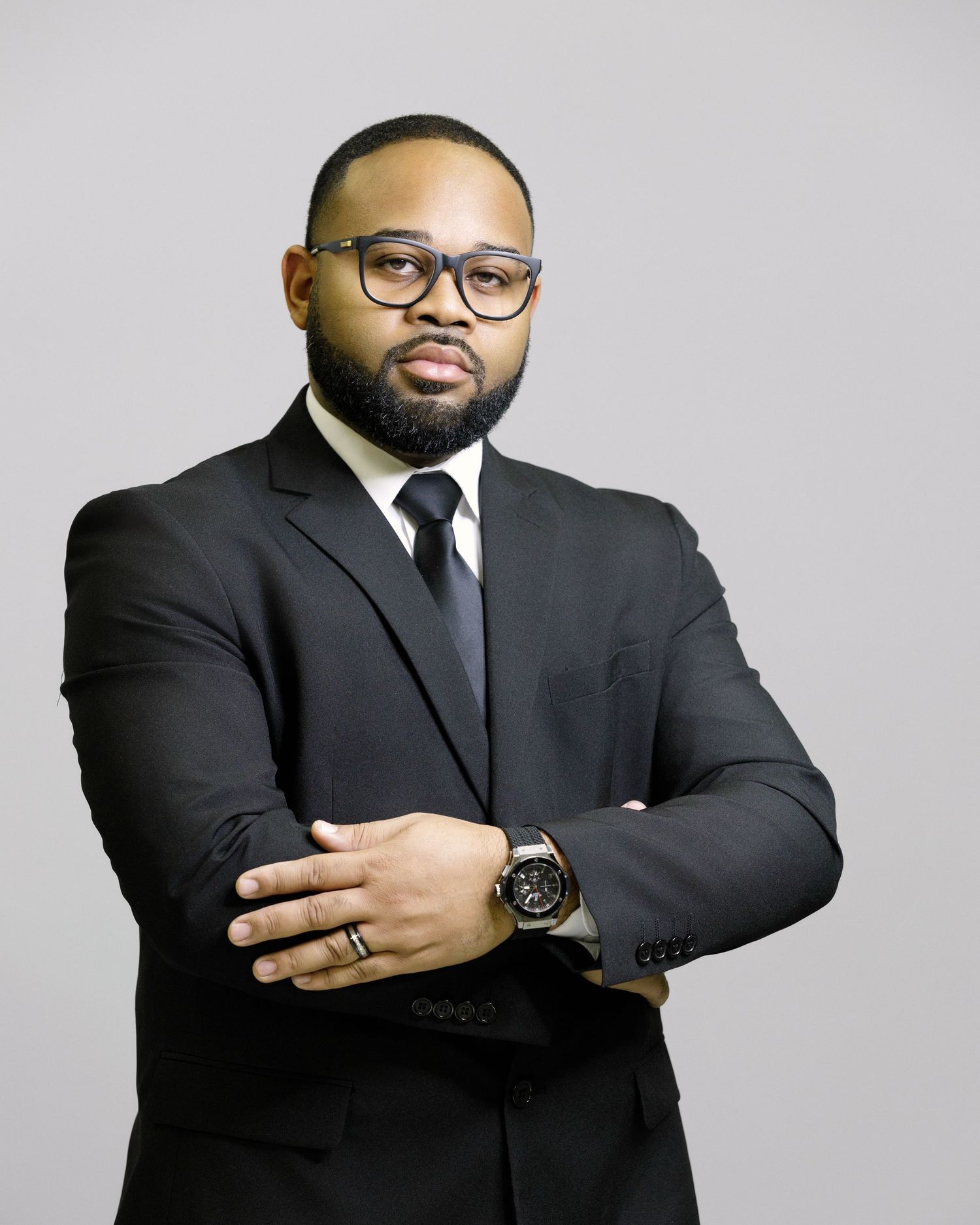 Professional headshots of man in black suit and glasses against light gray background.