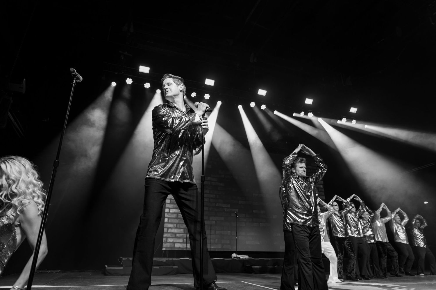 Black and white stage performance photo showing performers in dramatic lighting with spotlights and smoke effects.