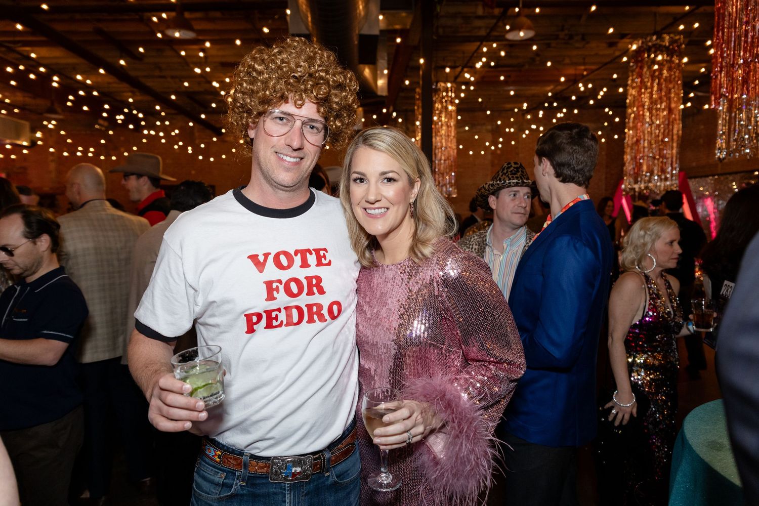 Two people pose at a party under string lights, with one wearing a 