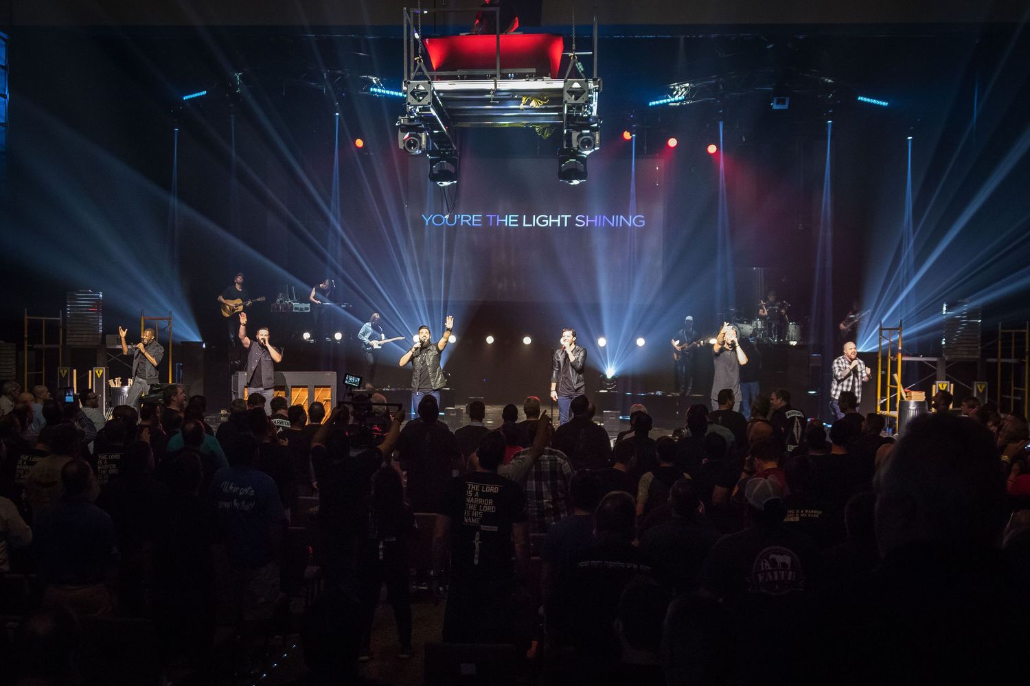 Concert performers on stage with dramatic blue lighting and beams illuminating a dark venue filled with spectators.