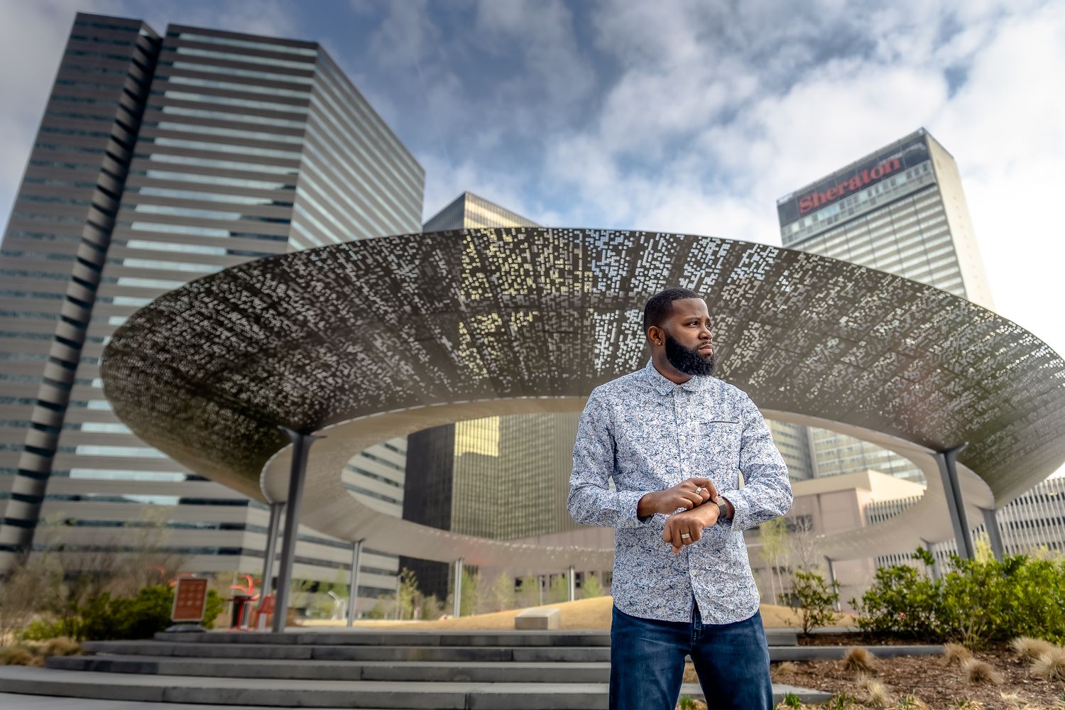 A person stands in front of a large metallic sculptural dish with modern office buildings in the background.