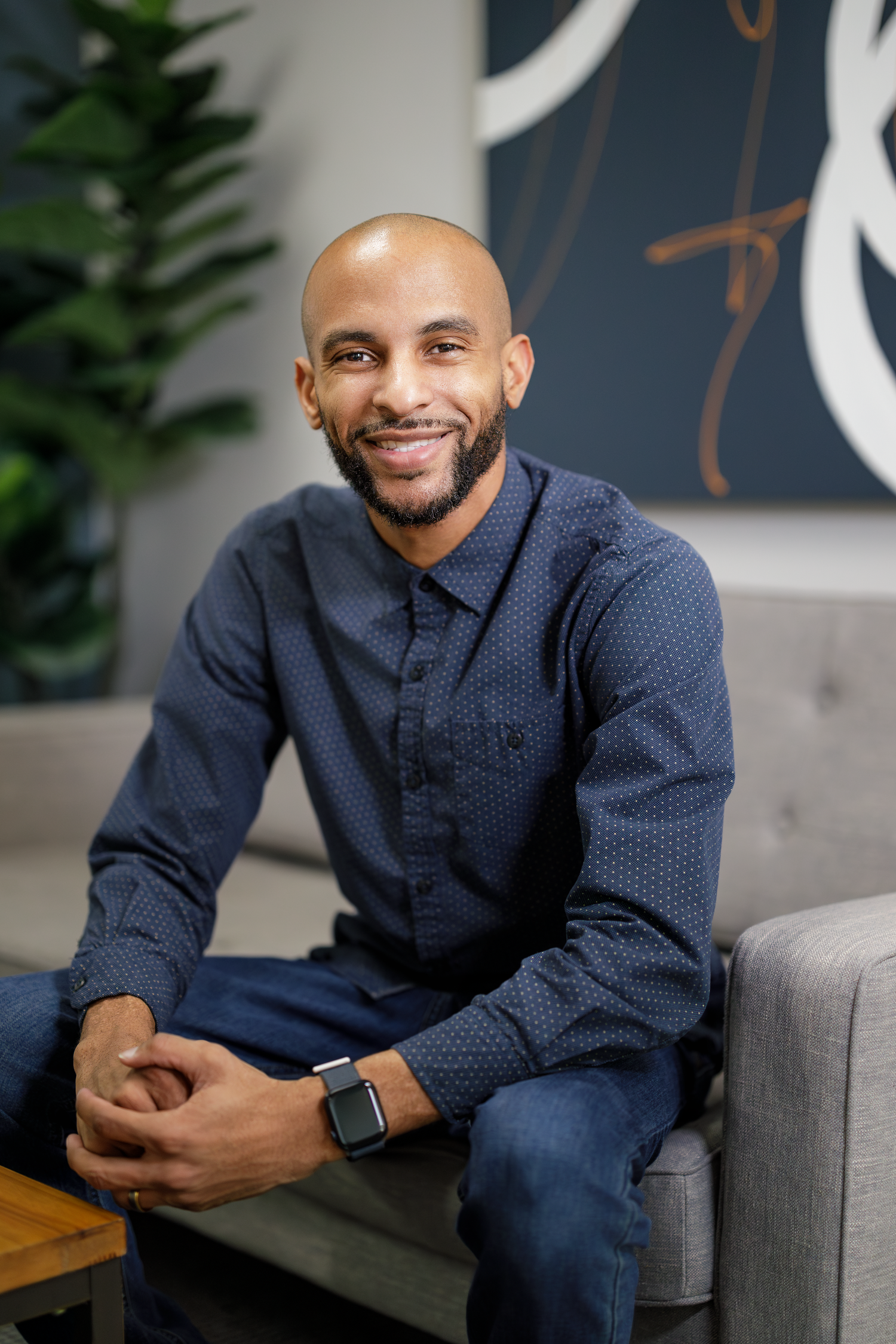 Professional headshots in navy blue shirt seated in grey armchair.