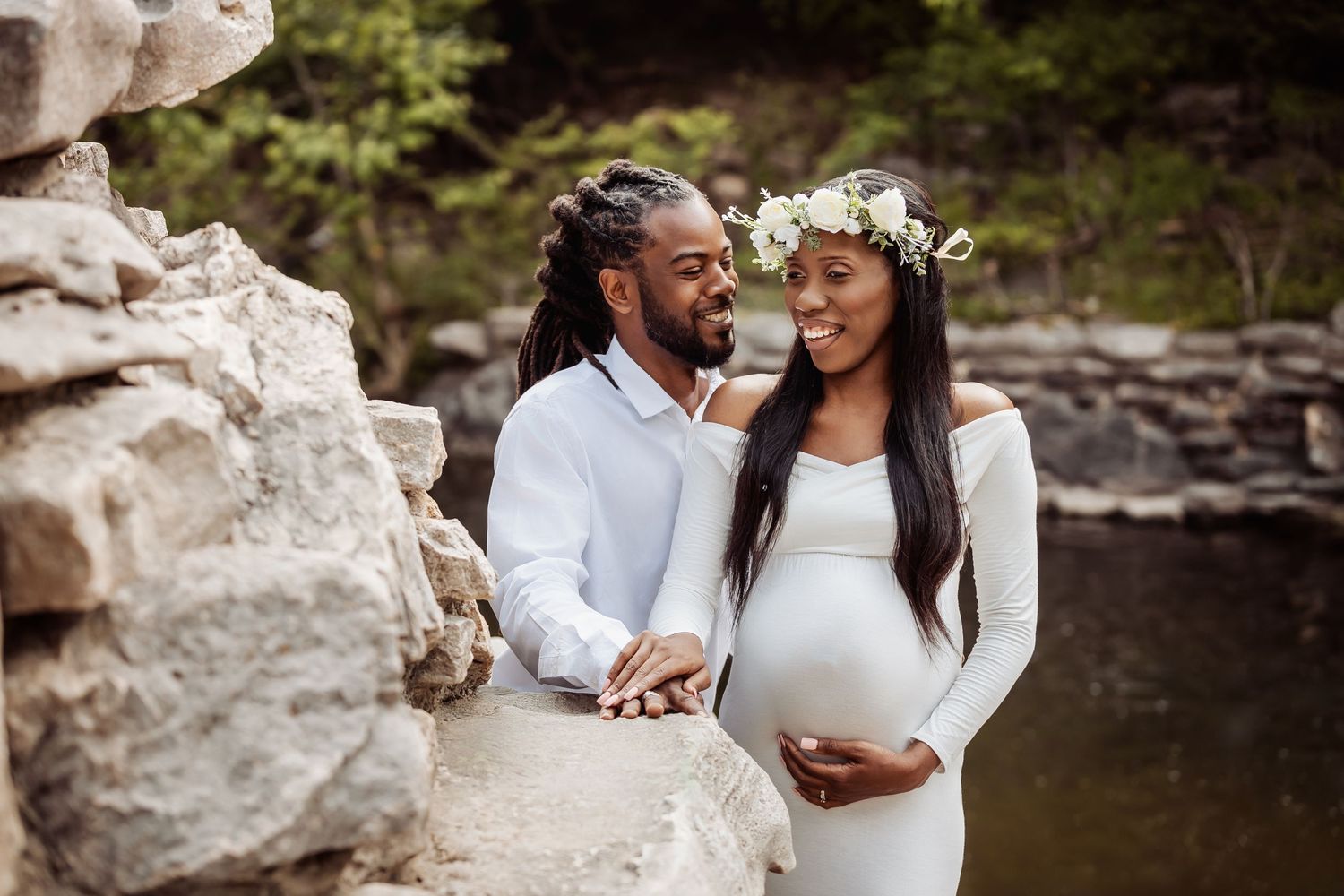 A couple in white outfits shares an intimate moment during their maternity photoshoot by a serene lake.