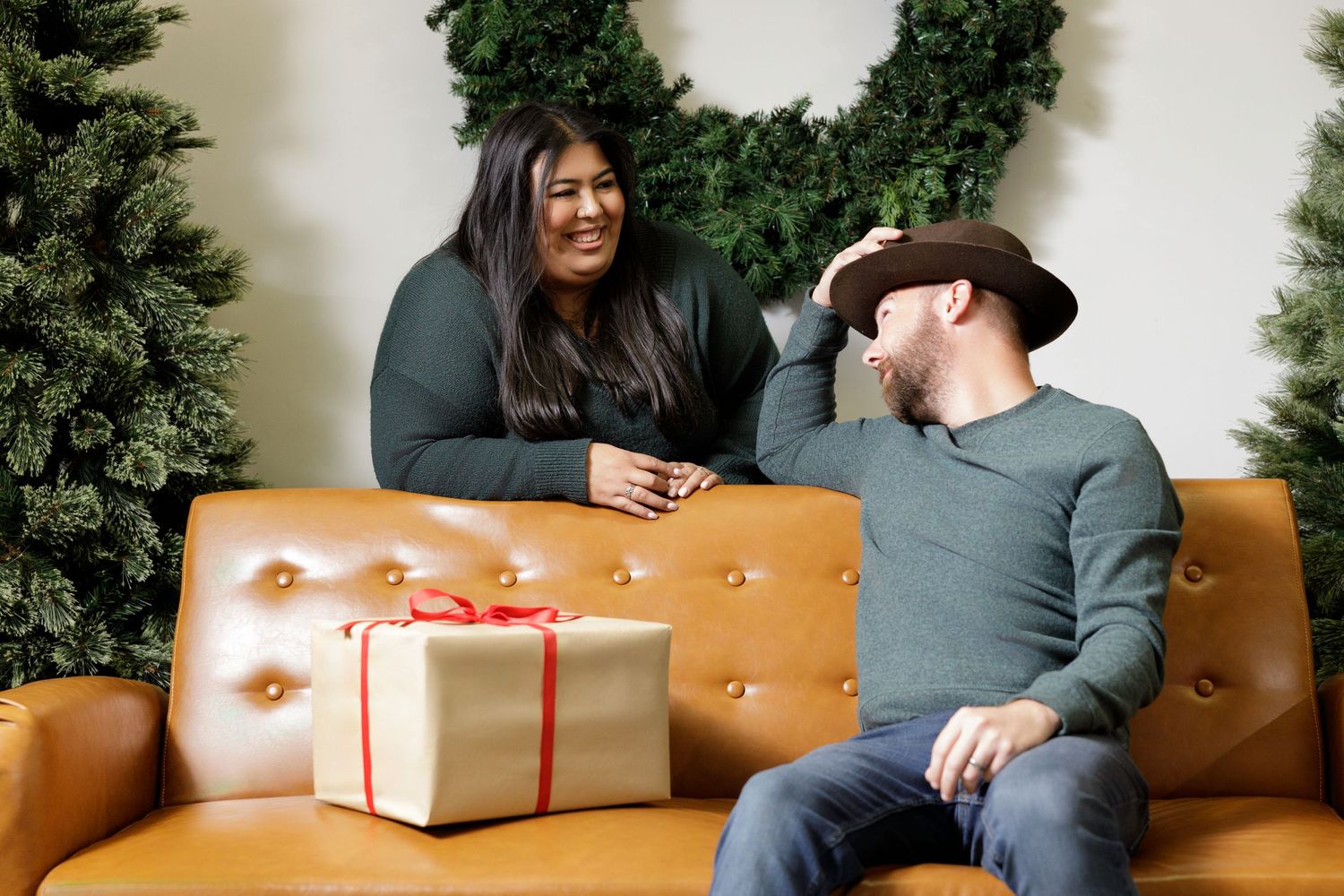 Holiday photoshoot setup with Christmas trees, presents, and leather furniture against white wall.