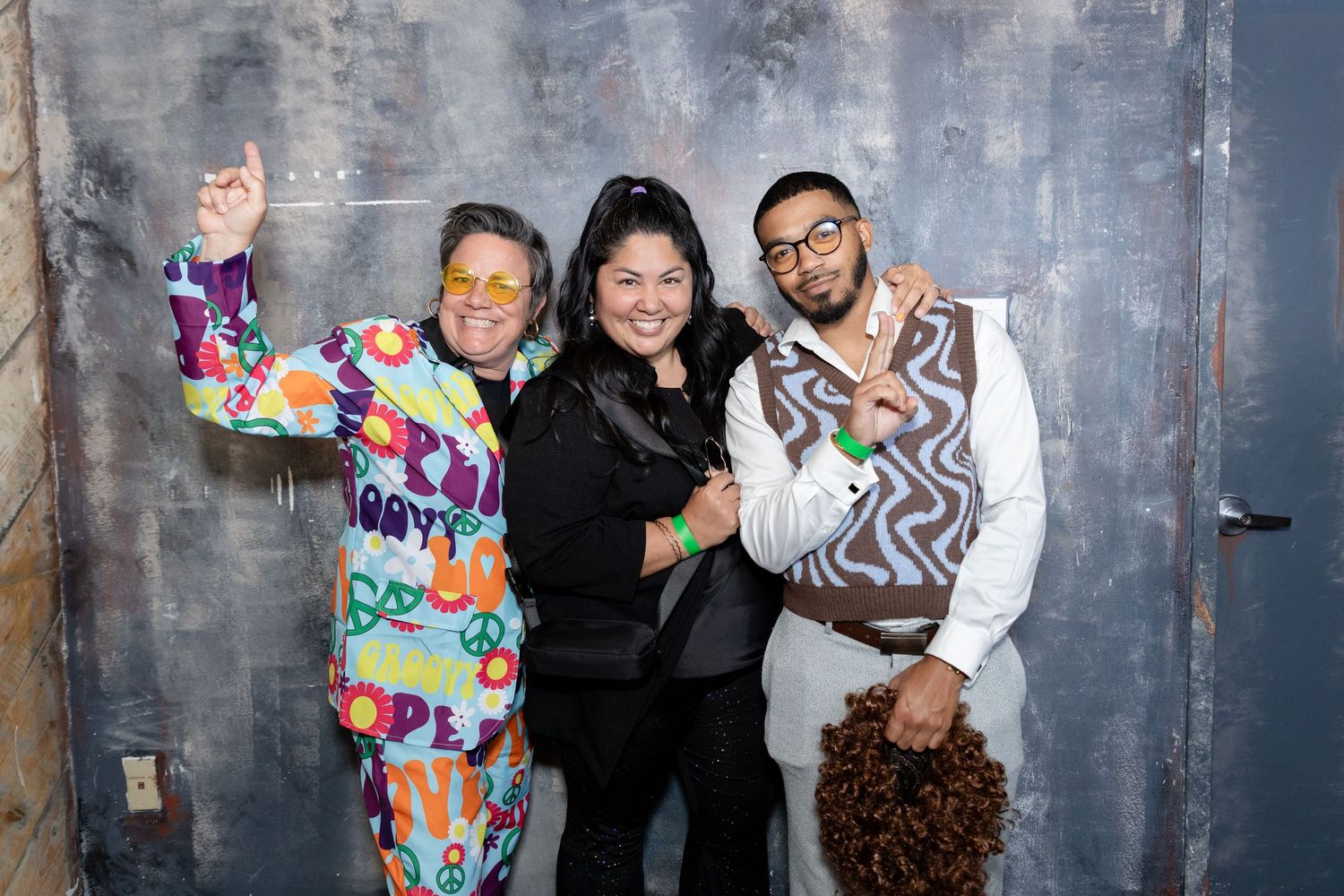 Three people pose together against a textured gray wall wearing colorful and casual outfits.
