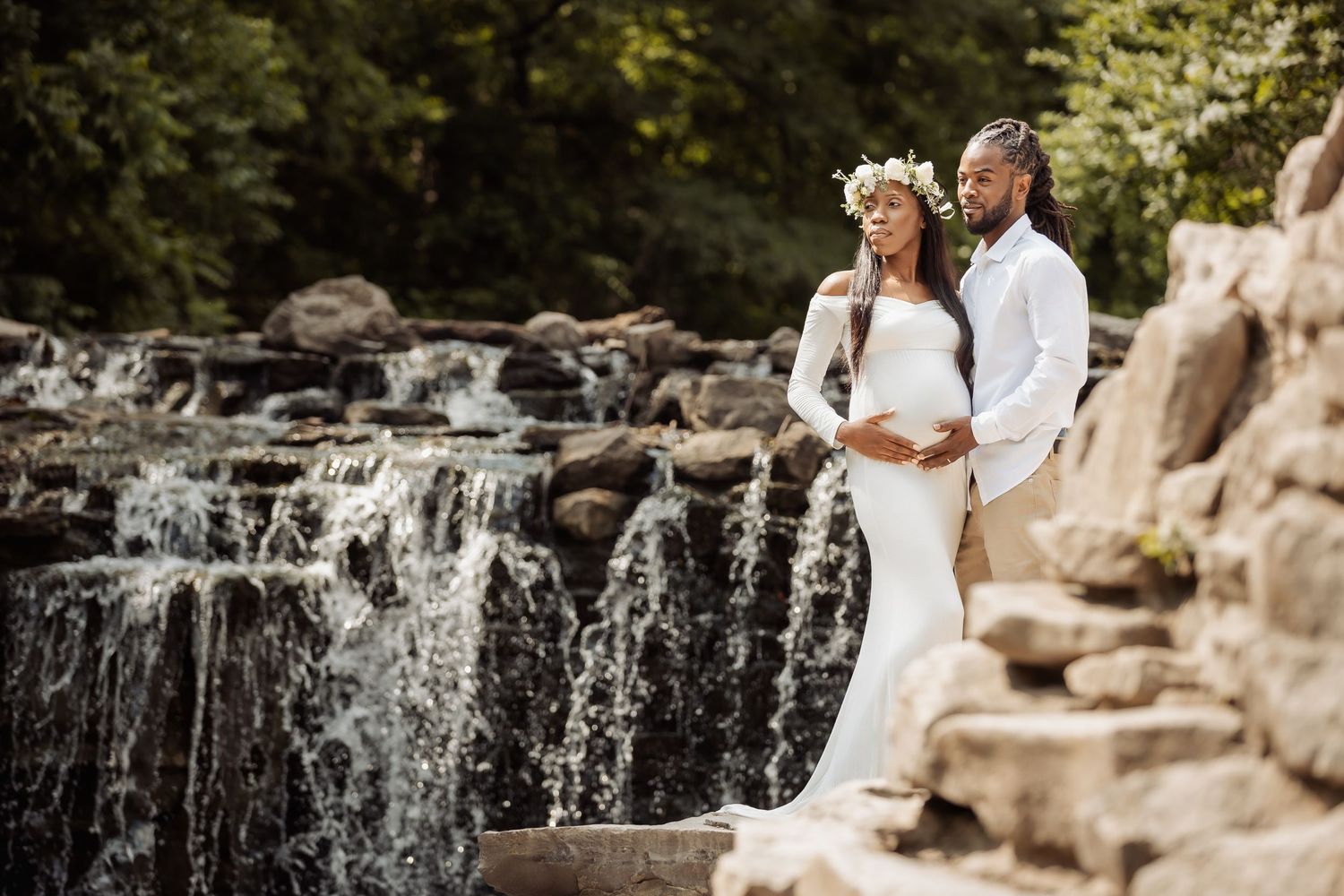 A couple in wedding attire embraces near a scenic waterfall with rocky outcrops in a natural outdoor setting.