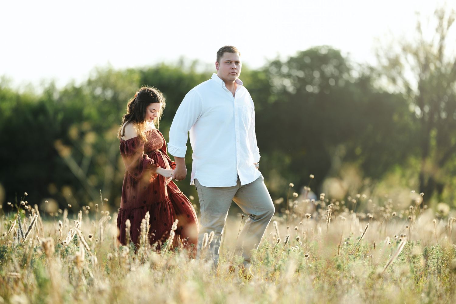 A couple walking through a golden sunlit meadow with tall grass and wildflowers at sunset.