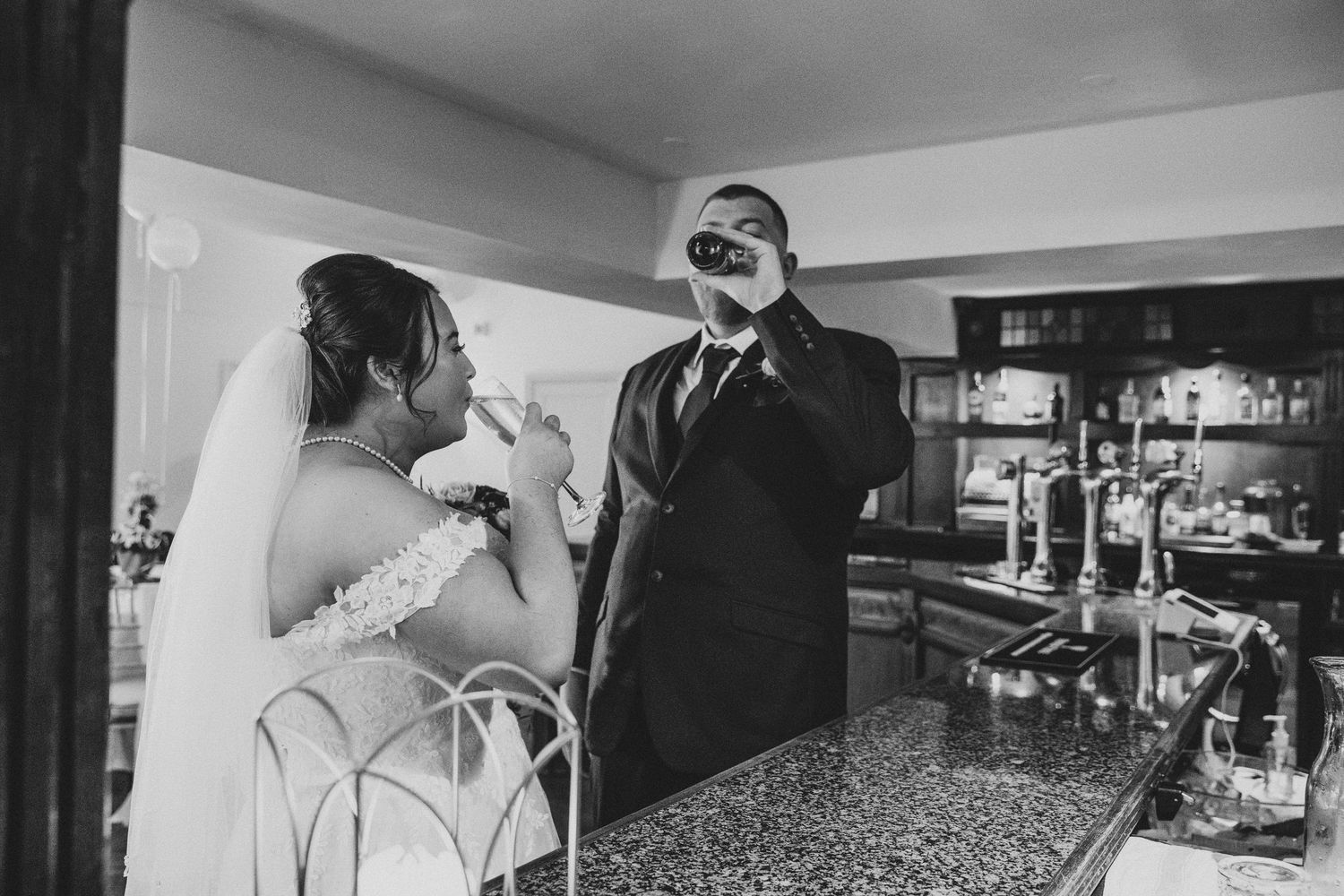 Bride and groom share a drink at the bar in a candid black and white wedding reception photo.