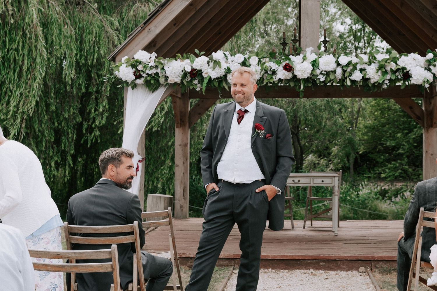Groom in gray suit with red tie smiling at outdoor wedding ceremony beneath floral arch.