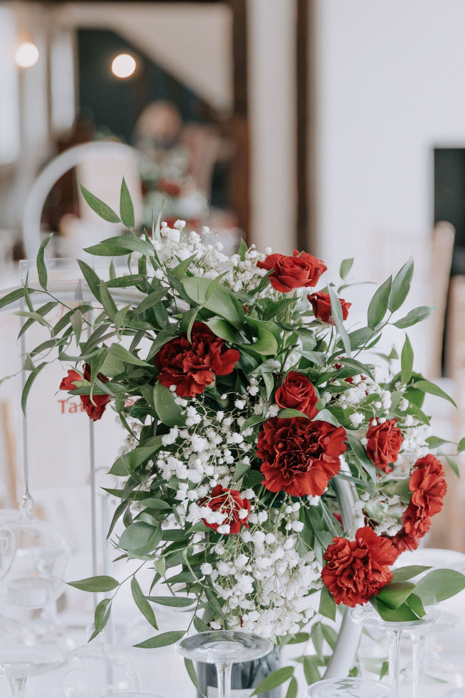 Elegant wedding centerpiece with red roses, carnations, baby's breath, and green eucalyptus in a glass vase on a white table.