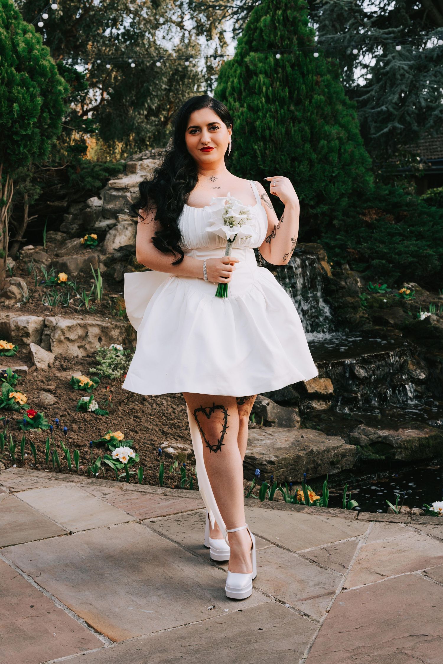 Woman in white mini dress holding bouquet poses in garden with waterfall backdrop.