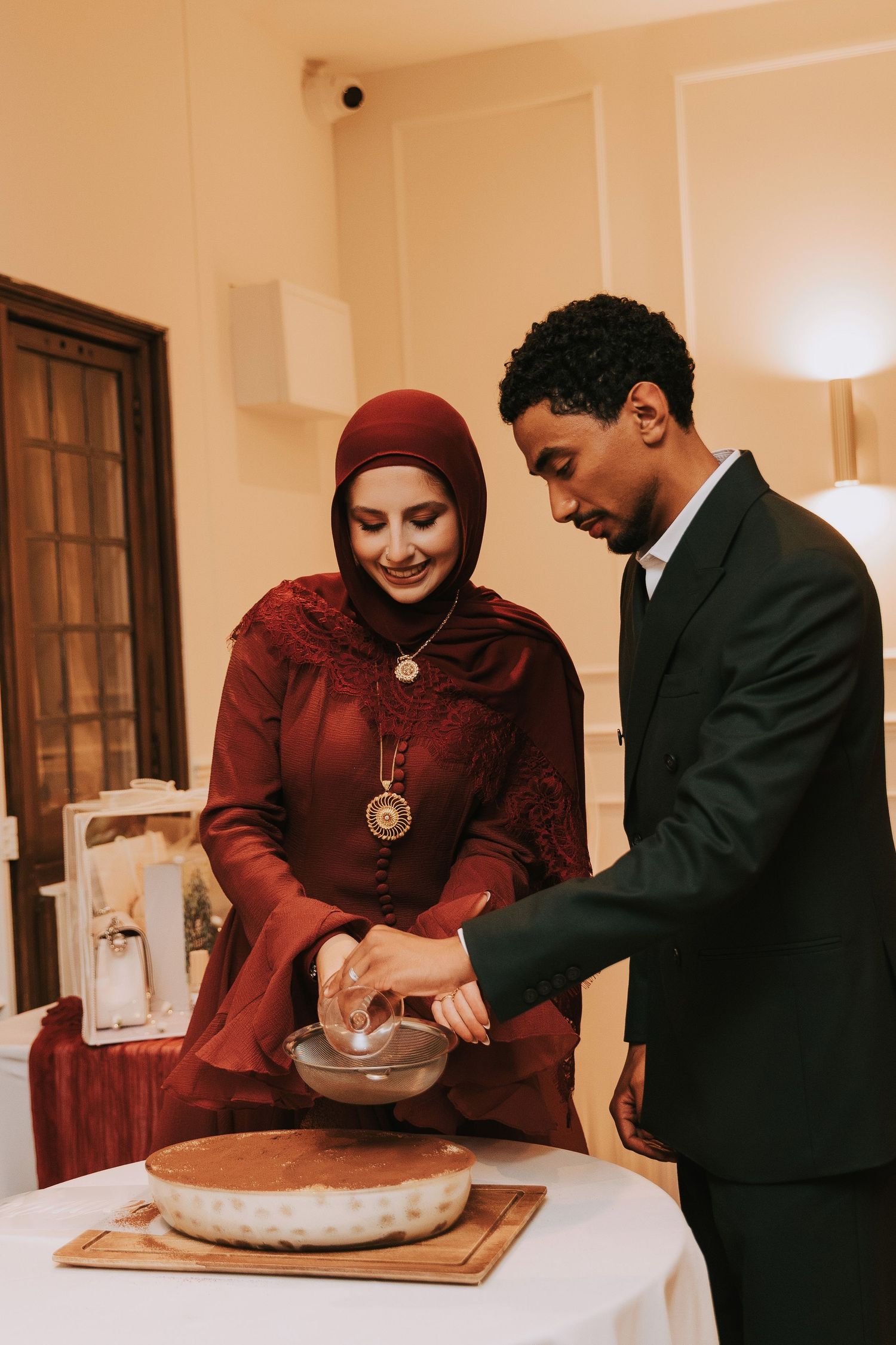 A couple in formal attire cutting a cake together at an elegant indoor celebration event.