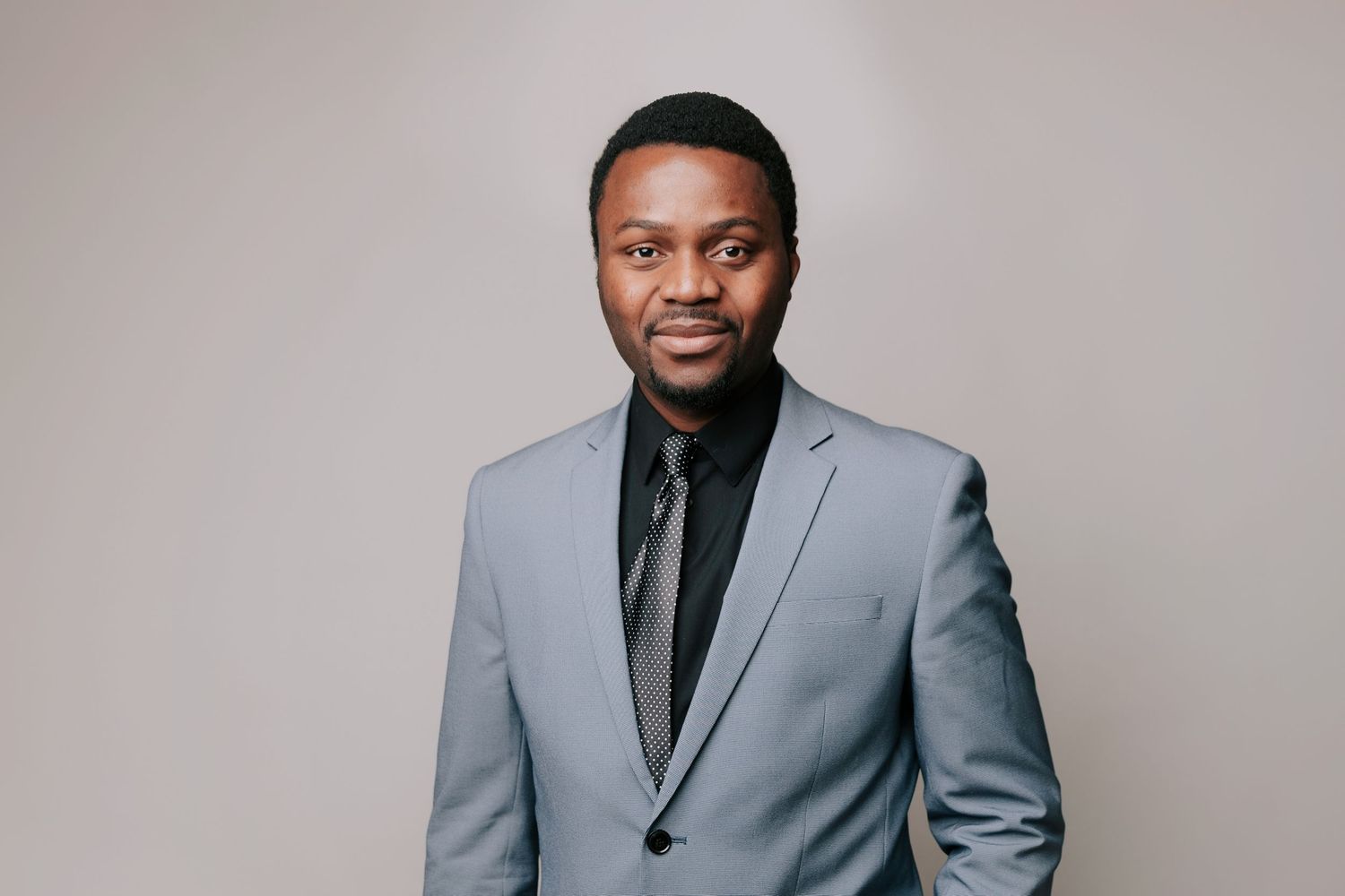 A man wearing a light grey suit stands in front of a plain grey color background, looking toward the camera ideal for professional headshot