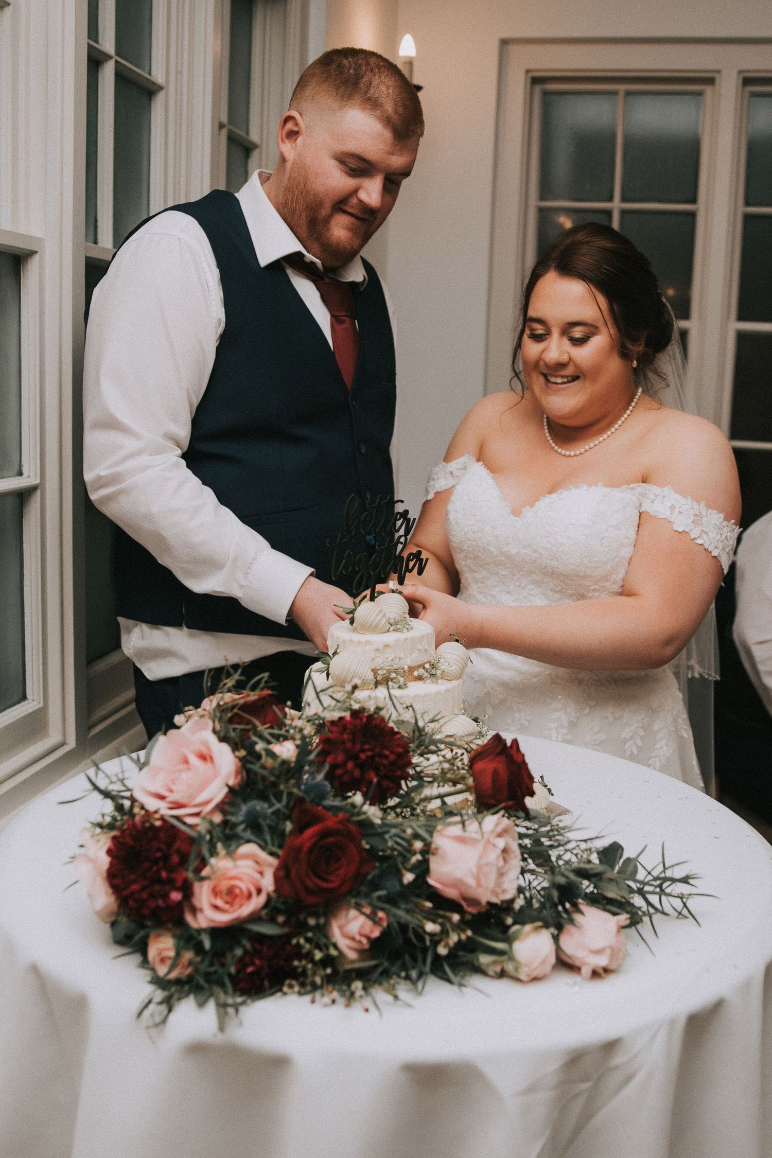 Smiling bride and groom cutting their white wedding cake surrounded by burgundy and pink roses.