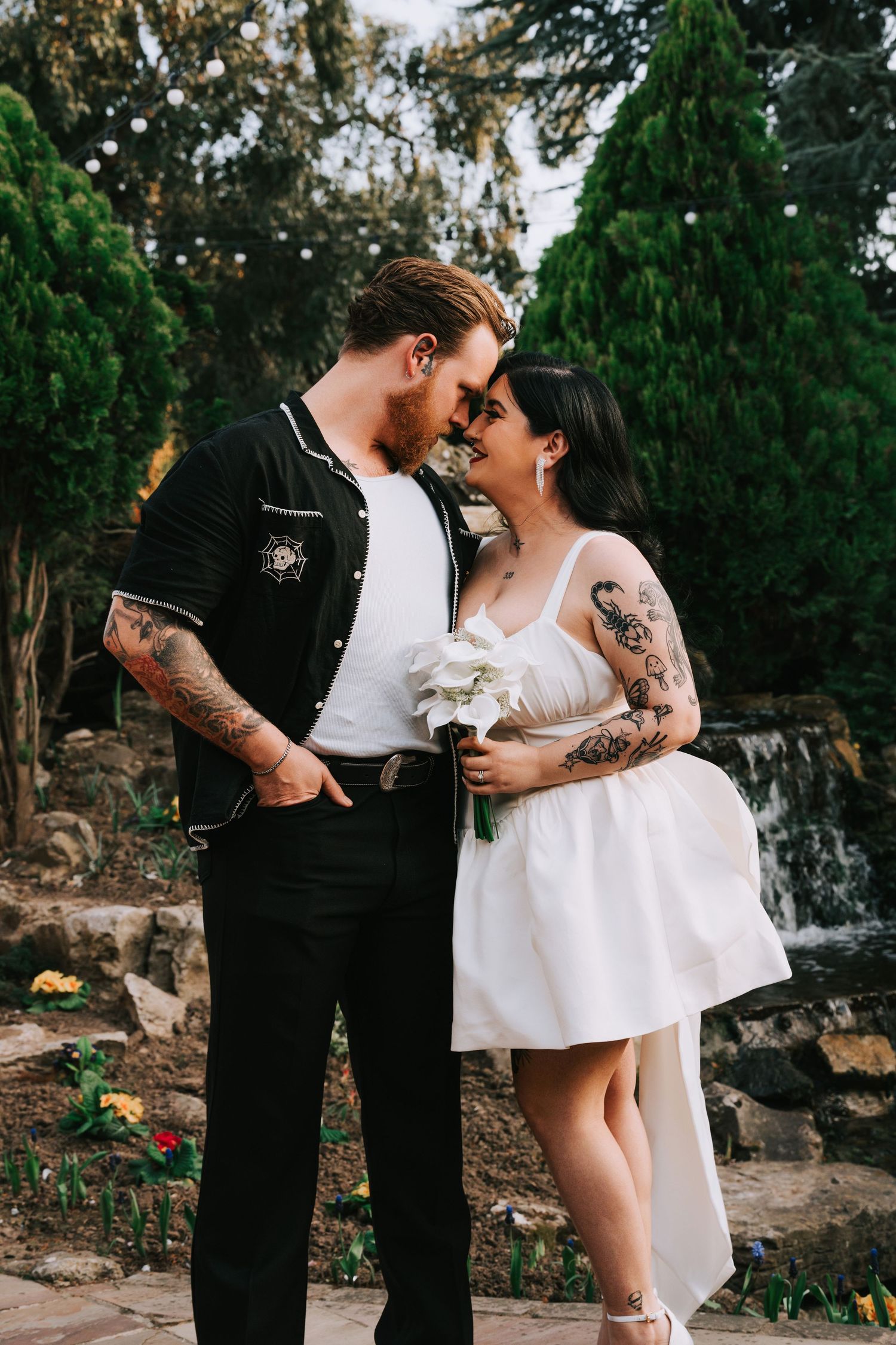 Tattooed couple in wedding attire sharing romantic moment outdoors near waterfall, bride holding white bouquet.