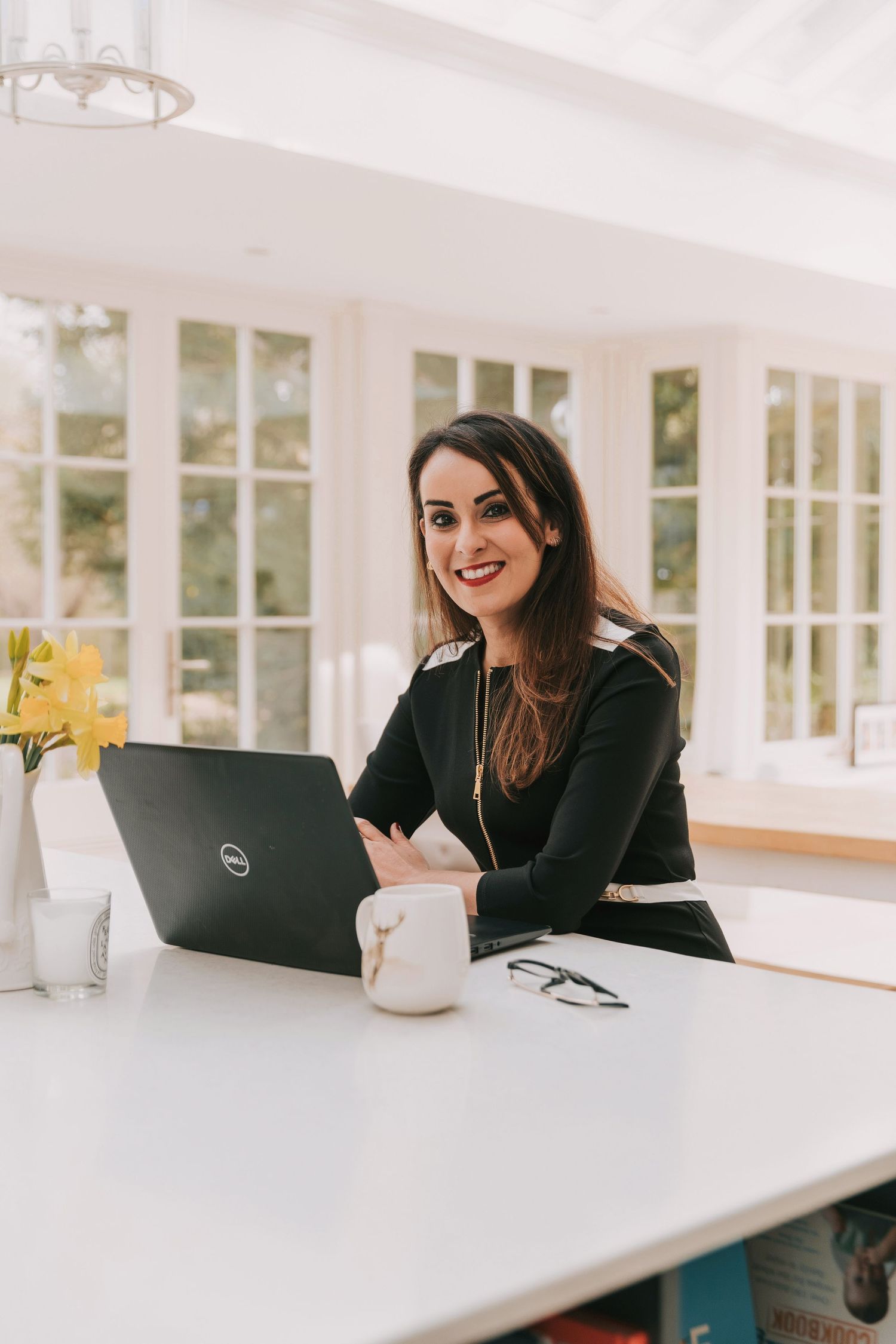 Professional headshot of a smiling GP & Coach in black suit dress working on laptop at white desk in bright home office in Basildon.