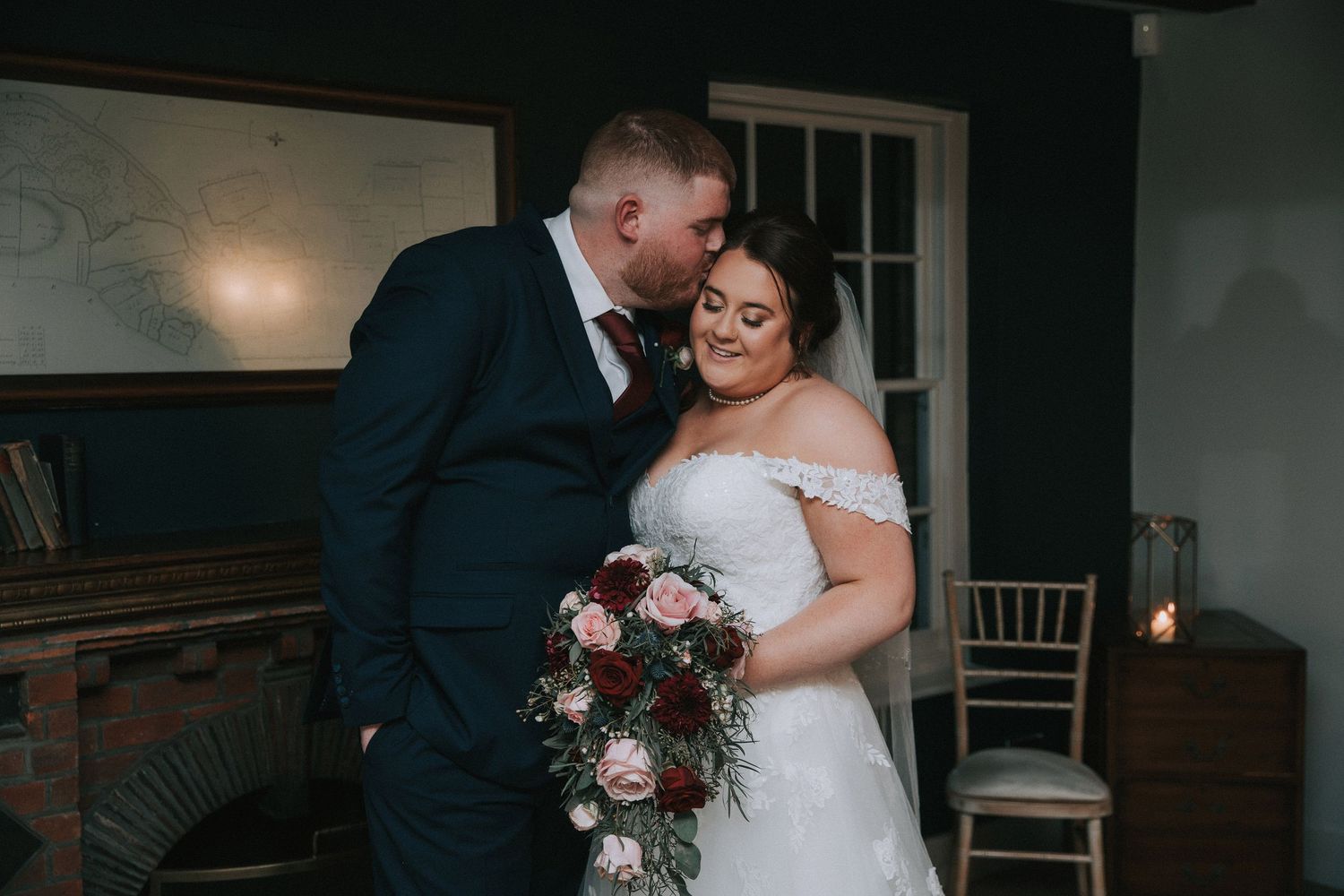 Groom in navy suit kissing bride in lace wedding dress while she holds floral bouquet indoors.
