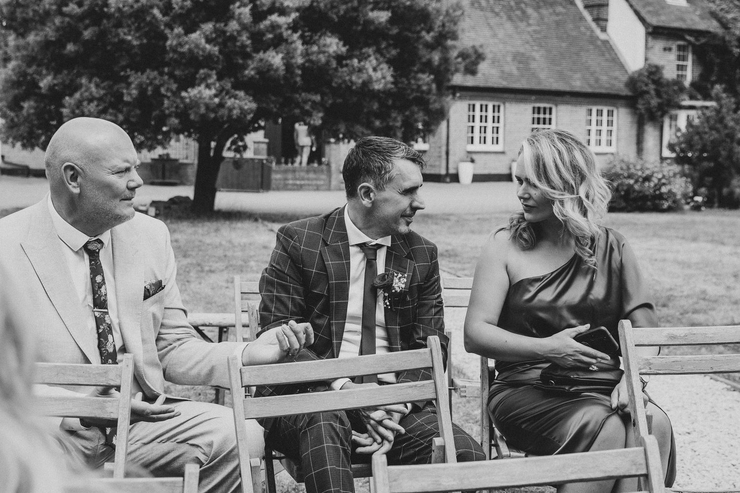 Black and white photo of two men and a woman laughing together on a wooden bench at an outdoor wedding venue.