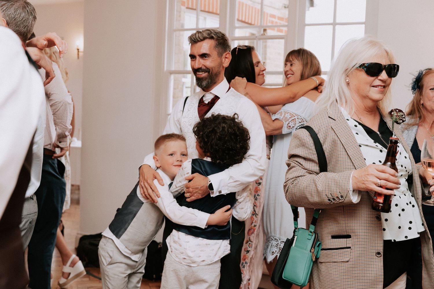 Man in suspenders hugging children at a wedding reception with guests socializing in background.