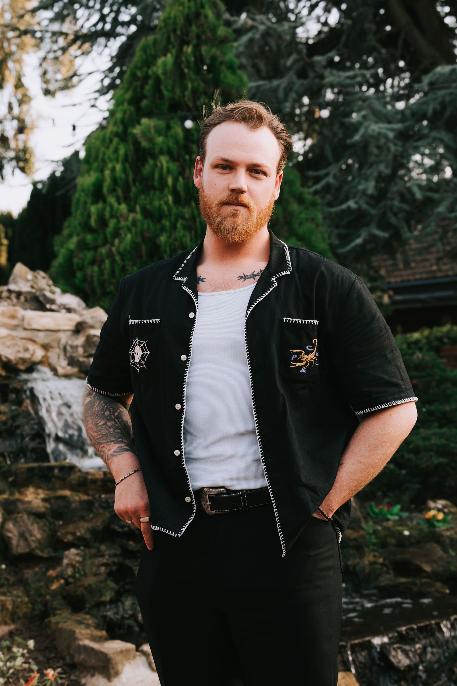 Tattooed bearded man in black embroidered shirt and white tank top posing outdoors near stone waterfall.