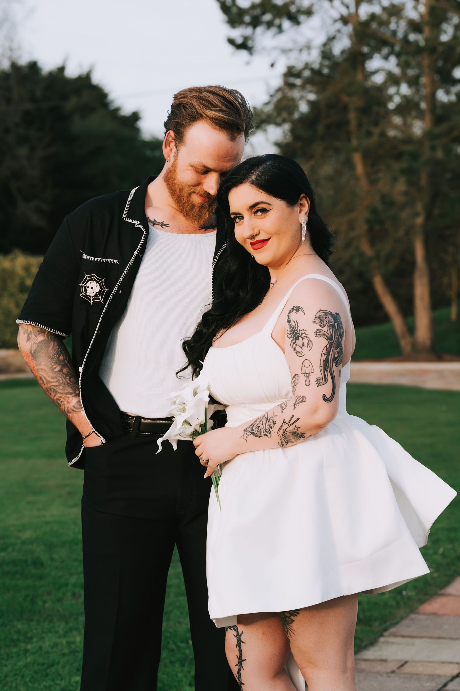 Tattooed couple in wedding attire pose outdoors, bride in white mini dress holds flowers, groom in black shirt smiles.