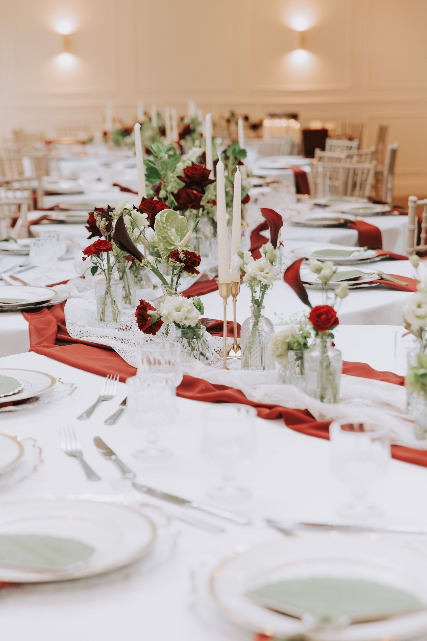 Elegant wedding reception table with red satin runners, white plates, crystal glasses, candles, and red and white floral centerpieces.