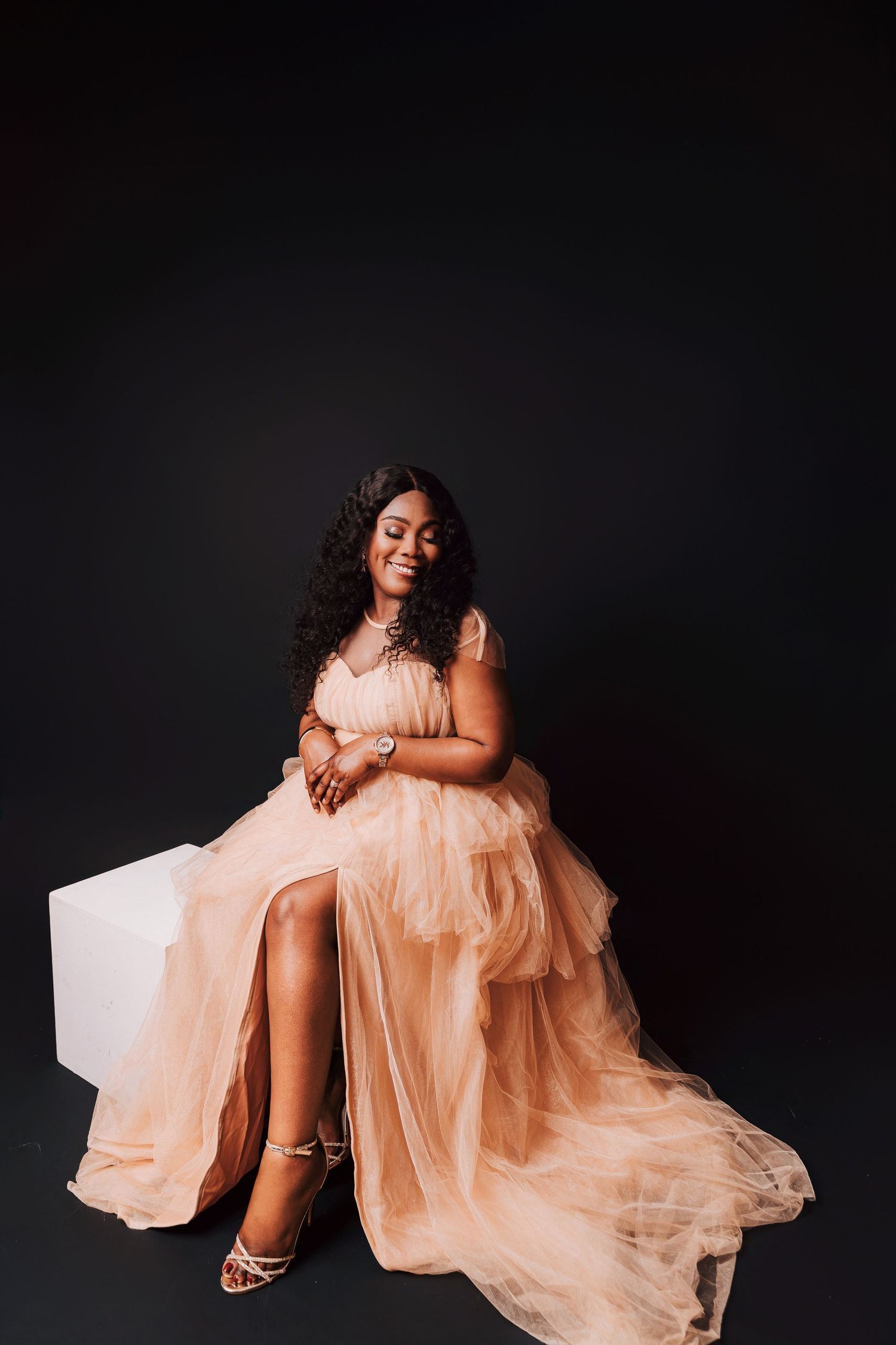 A woman in a peach tulle gown sits gracefully on a white cube against a black background, smiling softly.