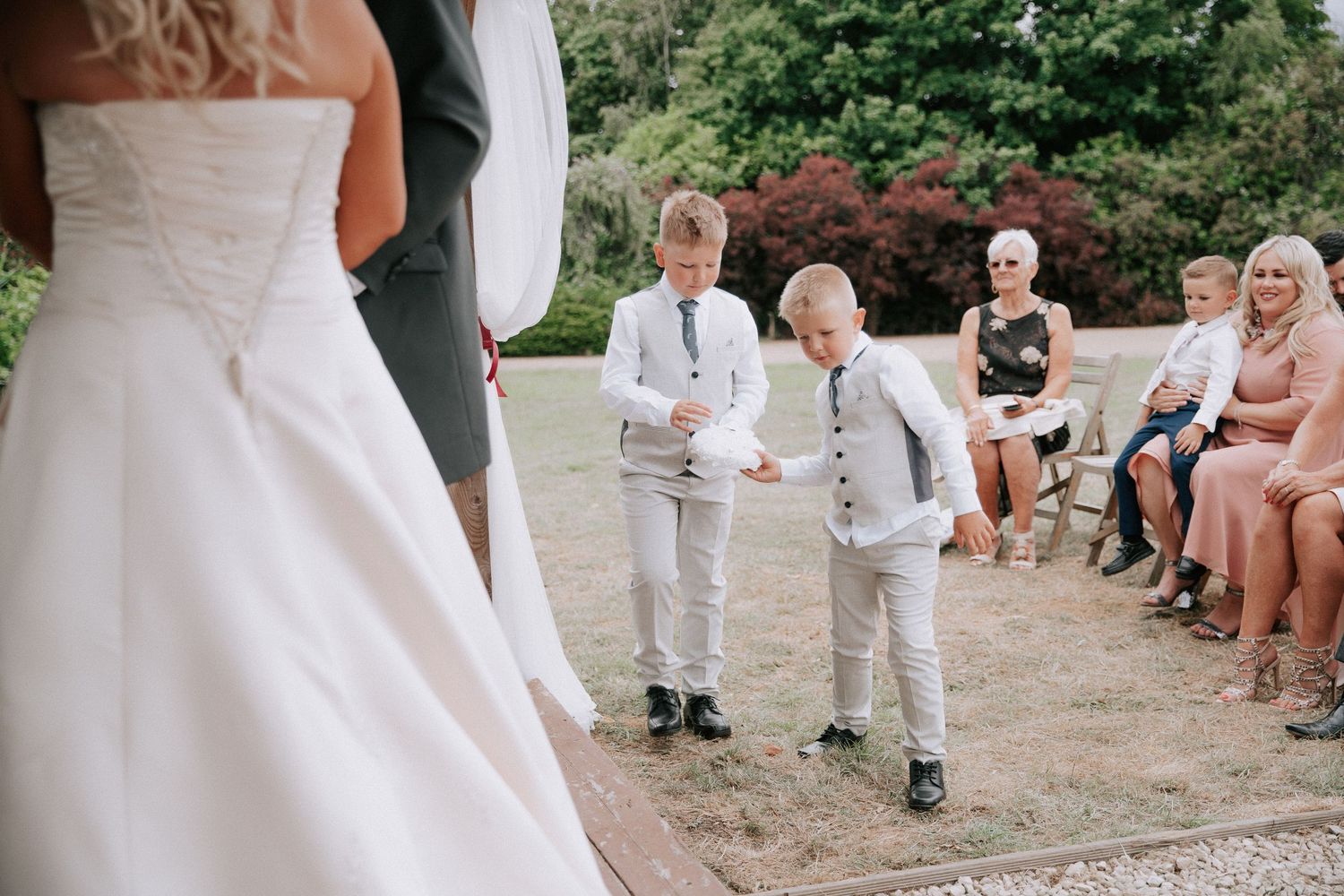 Two young ring bearers in grey suits present rings during an outdoor wedding ceremony with guests seated nearby.