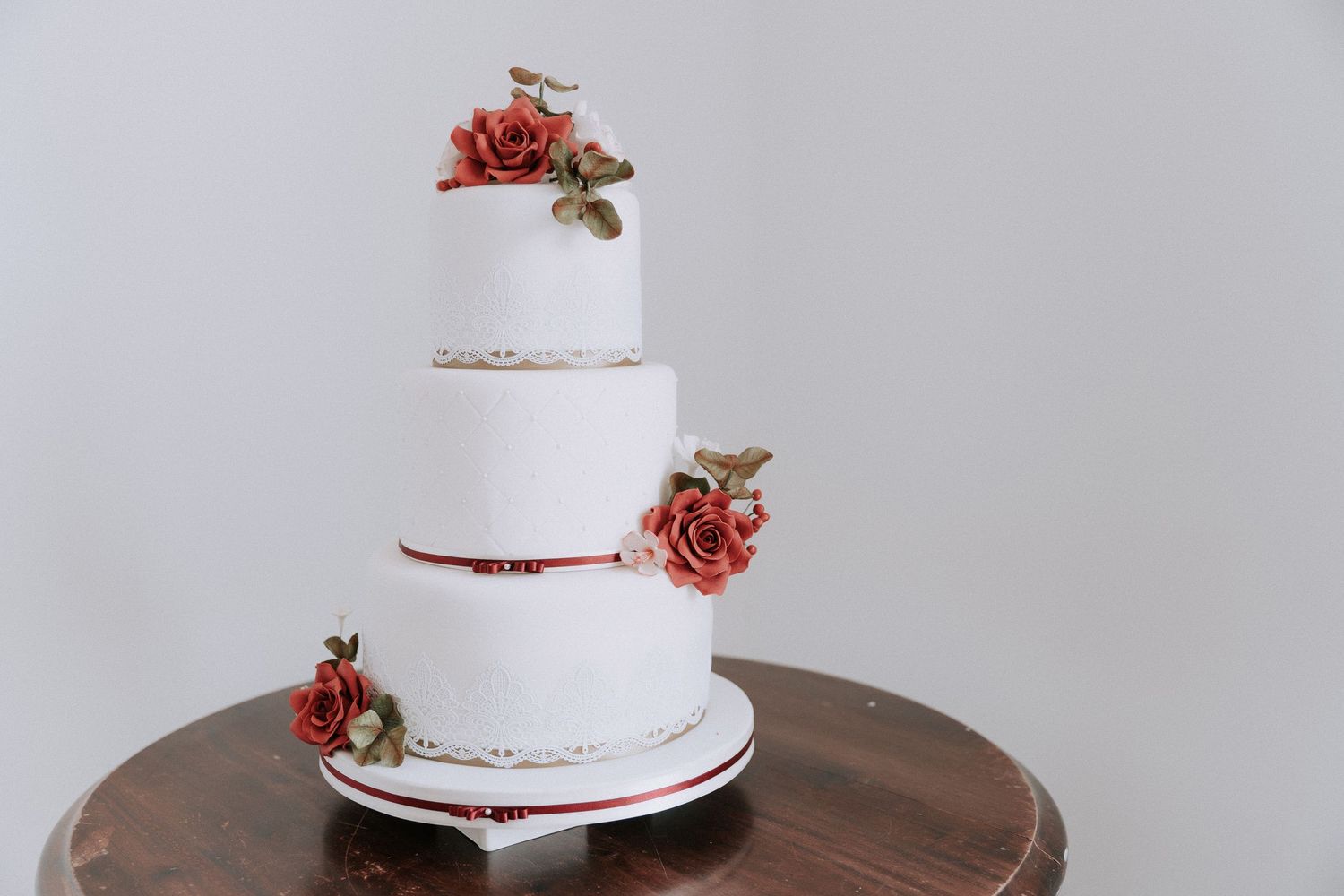 Cromwell Manor Basildon Wedding Cake Three-tier white wedding cake with red roses and gold leaf details on a wooden stand.