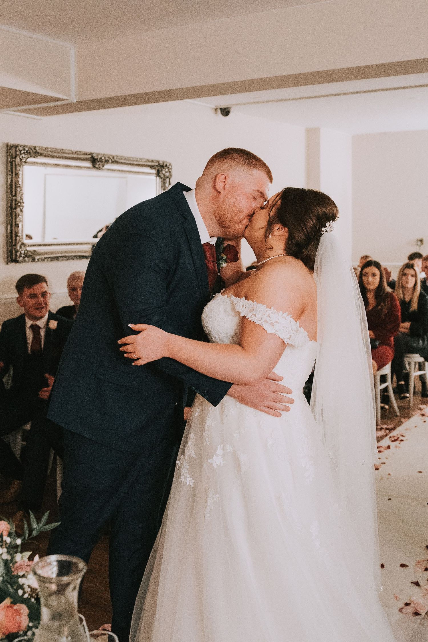 Bride and groom share their first kiss at the wedding ceremony altar, guests seated in the background.