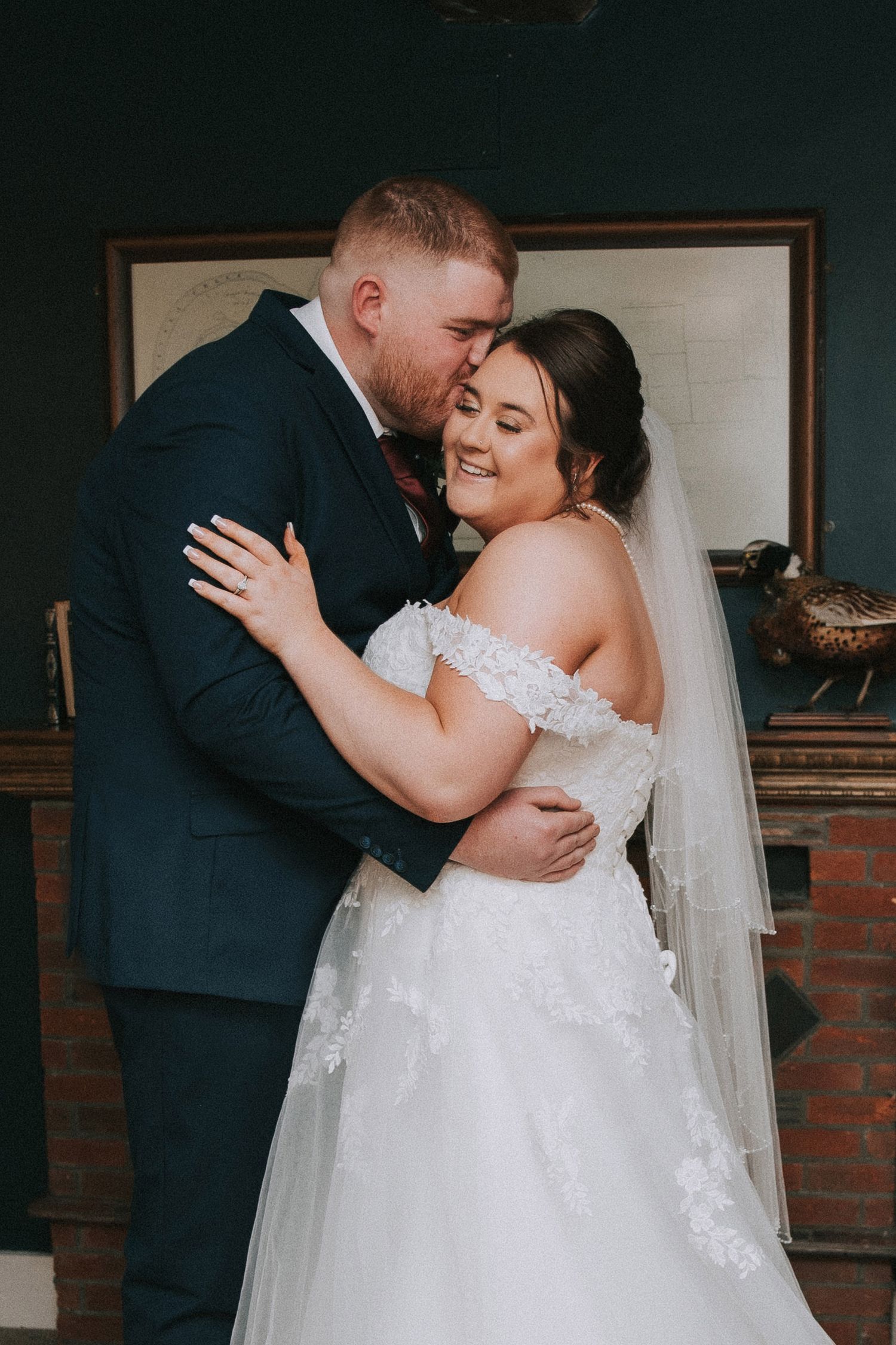 Groom in navy suit kissing bride on cheek, bride in lace off-shoulder wedding dress with veil, smiling warmly.