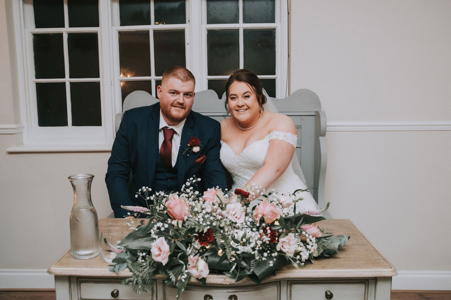 Happy newlyweds sitting together at a decorated table with pink floral arrangements on their wedding day.