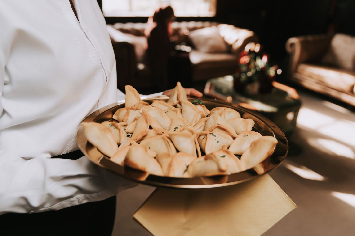 Waiter in white shirt holding a round silver tray of golden dumplings or pierogi at a catering event.