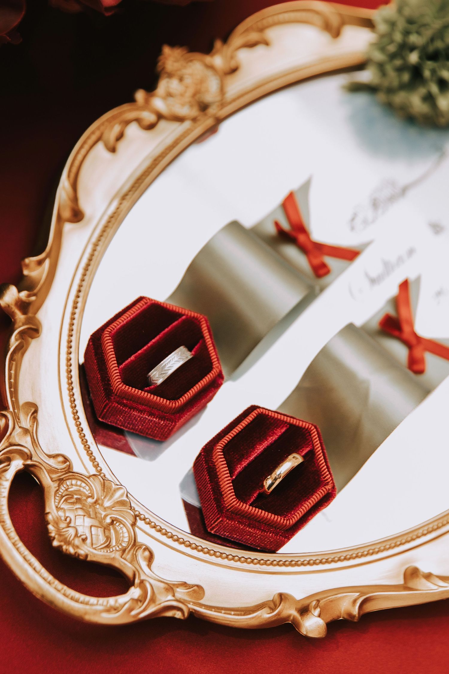 Two gold wedding rings displayed in red velvet hexagonal boxes on an ornate gold mirror tray with scroll invitations.