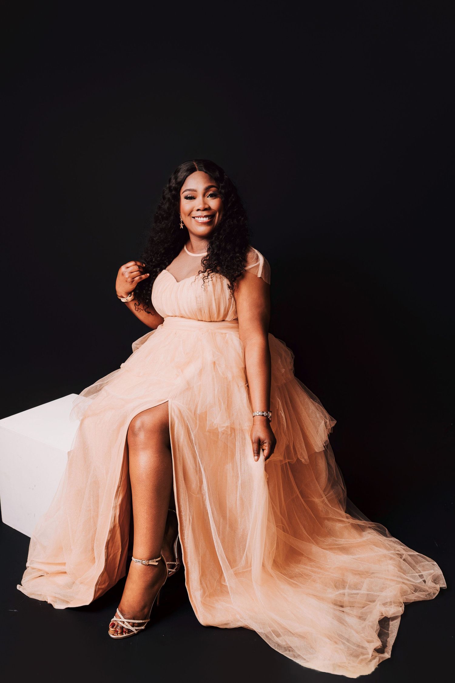 A smiling woman in a elegant peach tulle gown with a thigh-high slit poses gracefully against a black background.