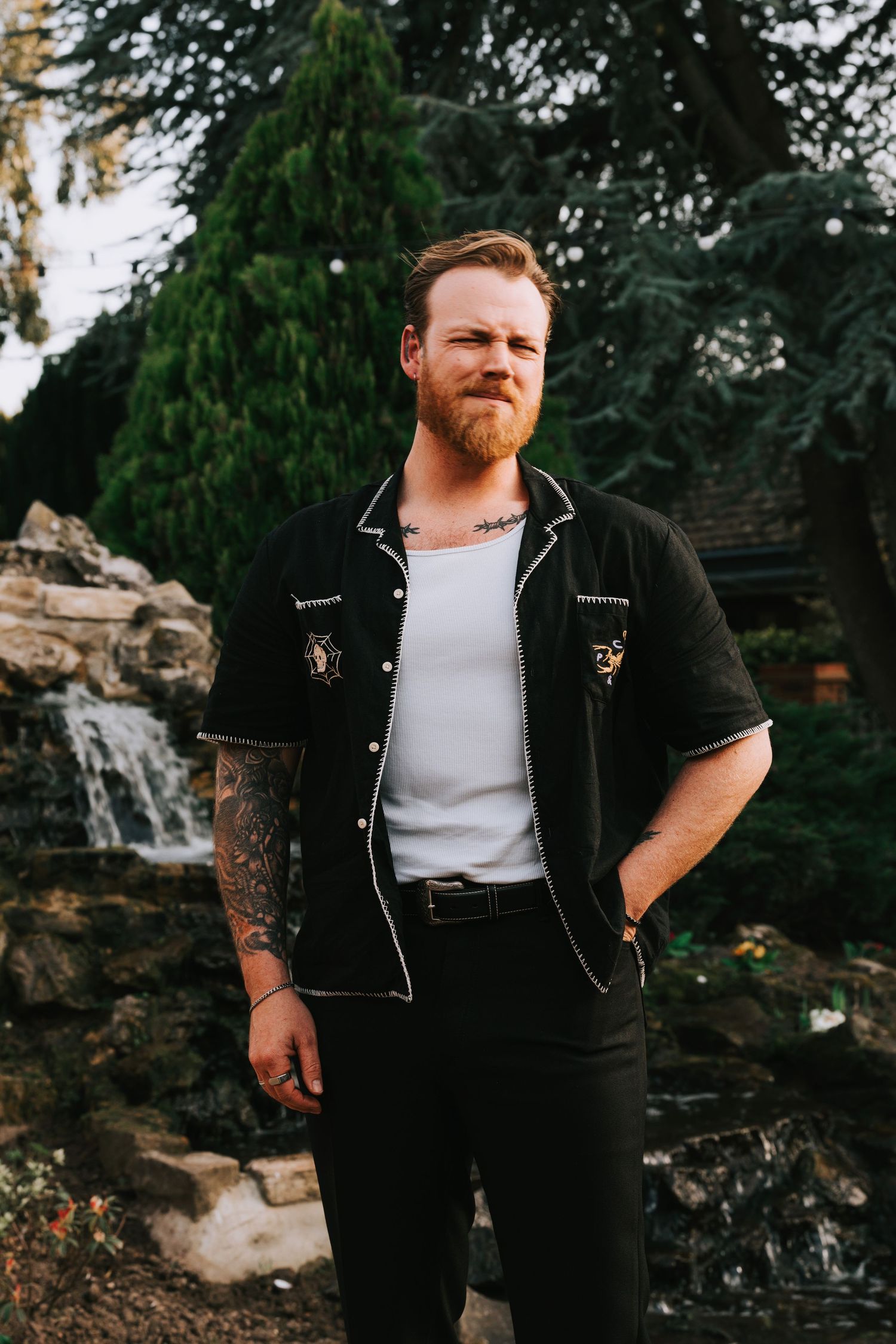 Tattooed redhead man with beard wearing black bowling shirt over white tank top, standing outdoors near waterfall.