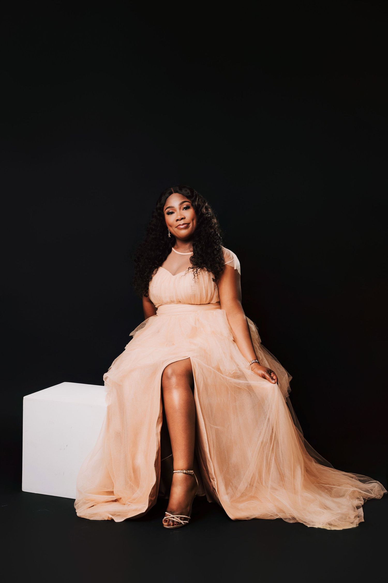 A woman in a peach tulle gown sits gracefully on a white cube against a black background.