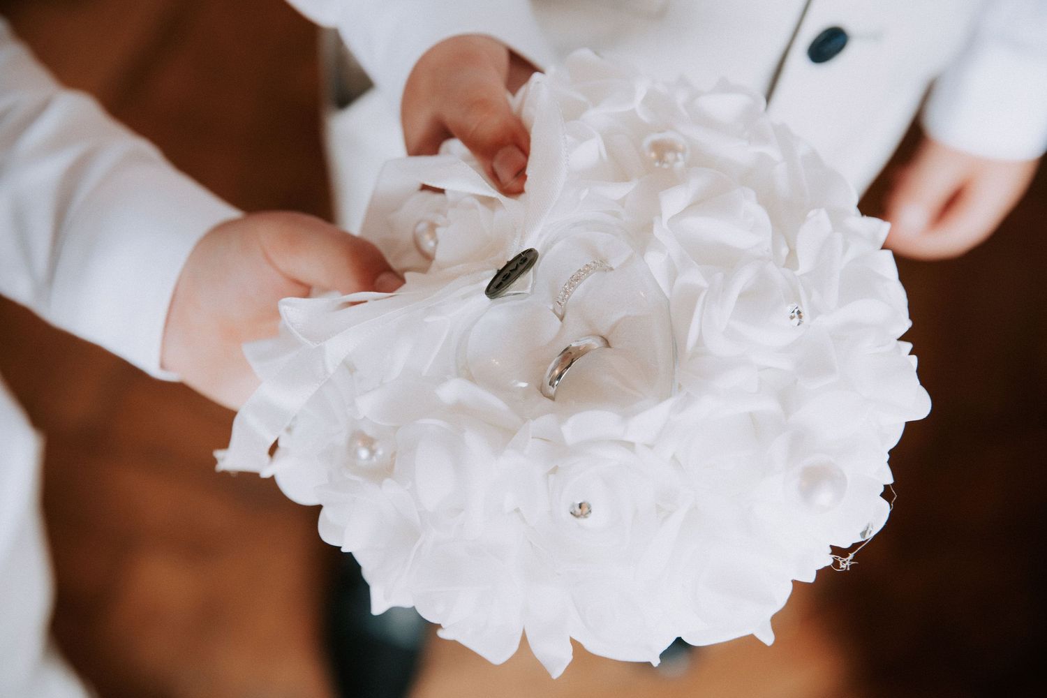 Child holding a white floral wedding ring pillow bouquet with pearl accents and wedding bands on top.
