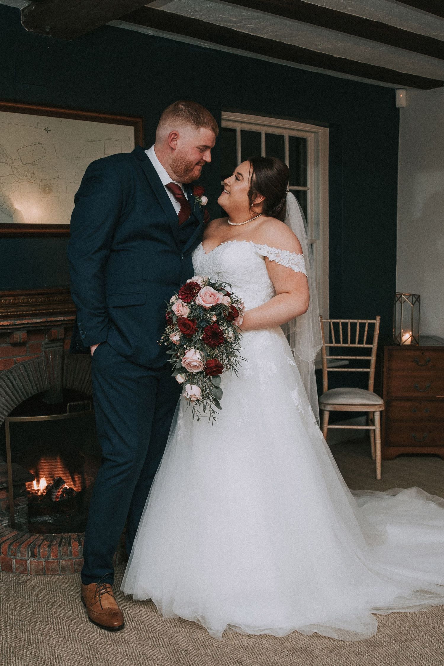 Bride and groom share a romantic moment by a fireplace, bride holding a burgundy and pink bouquet.