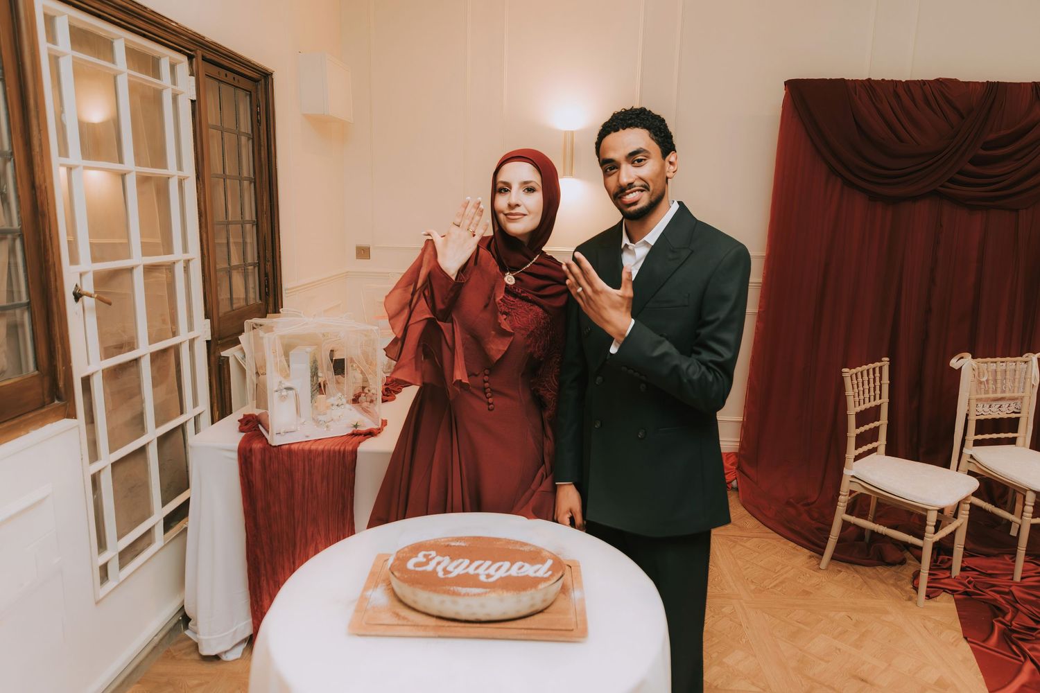 Muslim couple posing with an engagement cake at their engagement party, woman in burgundy hijab and dress.