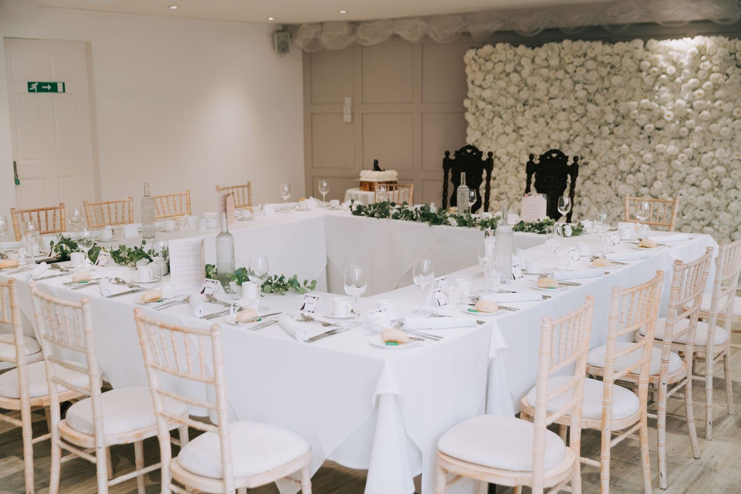Elegant wedding reception room with white tablecloths, chiavari chairs, floral wall backdrop, and greenery garland centerpieces.