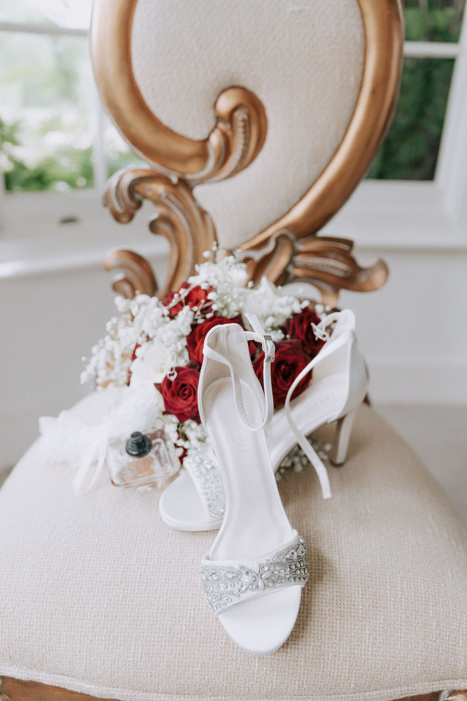 White bridal heels with crystal embellishments beside red roses bouquet on an ornate gold chair.