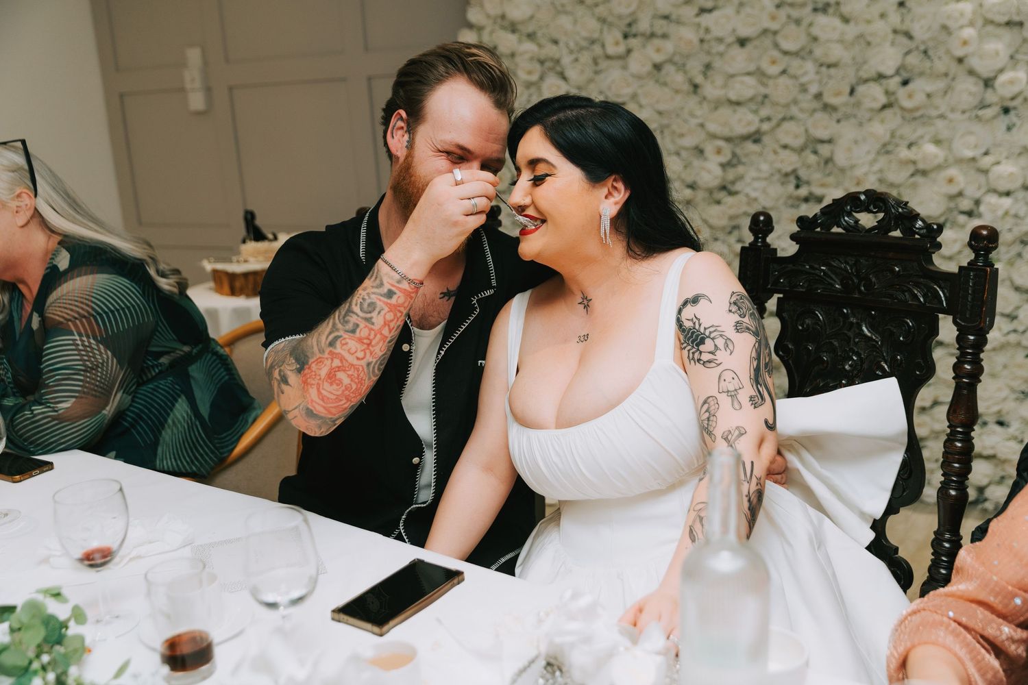 Tattooed bride and groom share an intimate laughing moment at their wedding reception table.