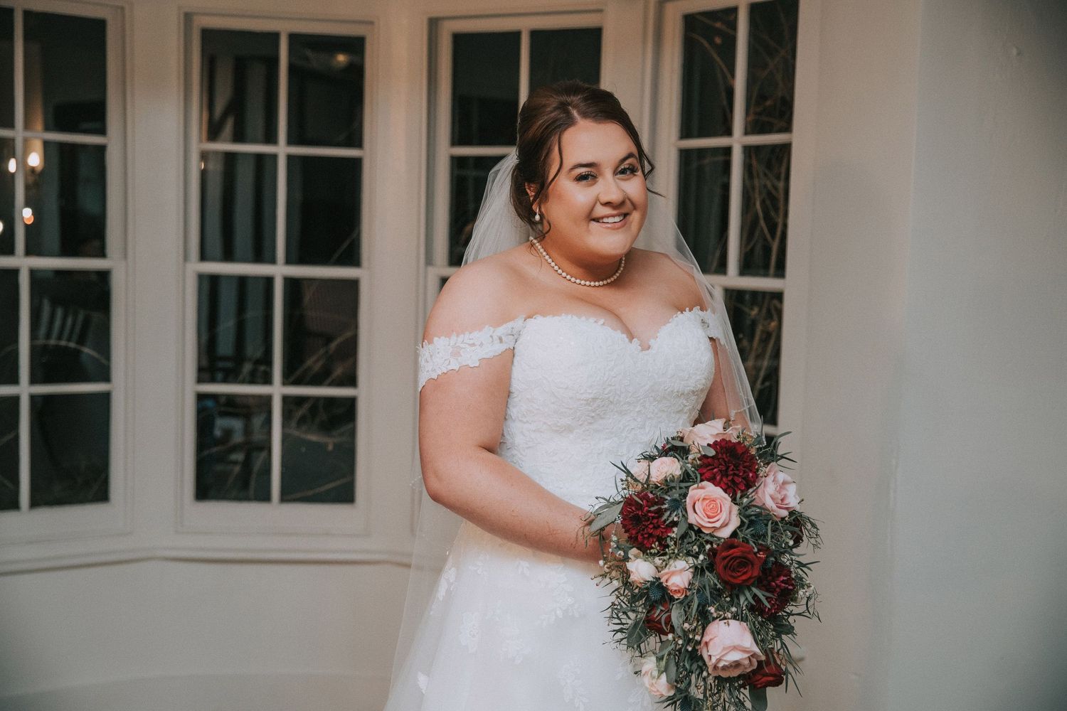 Smiling bride in off-shoulder lace wedding dress holding burgundy and pink rose bouquet by window.