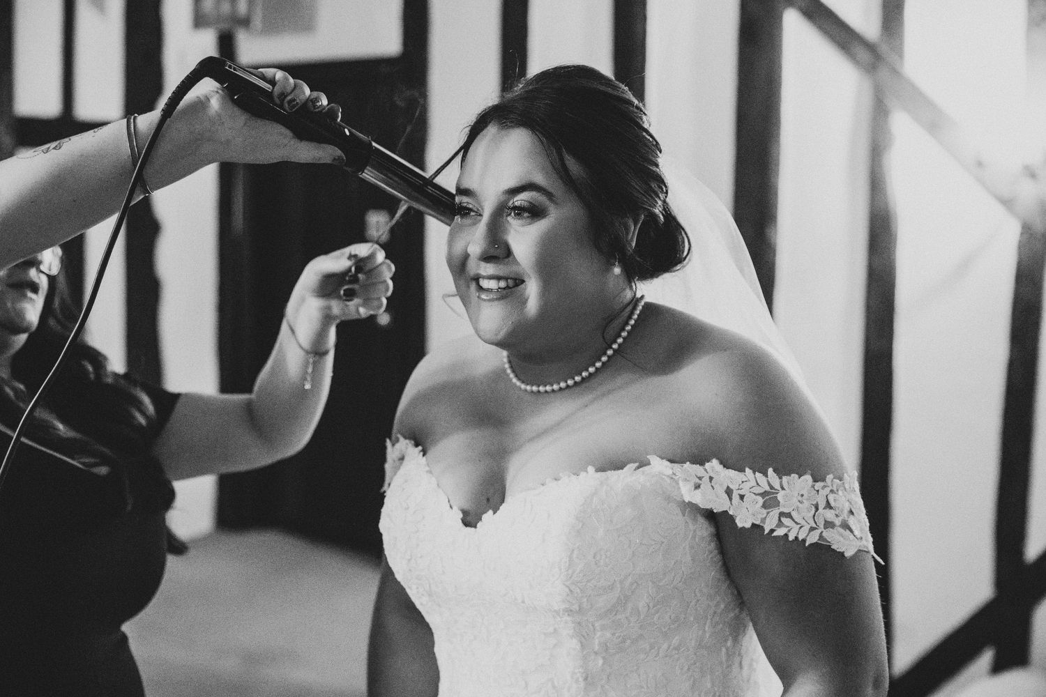 Black and white photo of bride in lace wedding dress getting hair styled with curling iron before ceremony.
