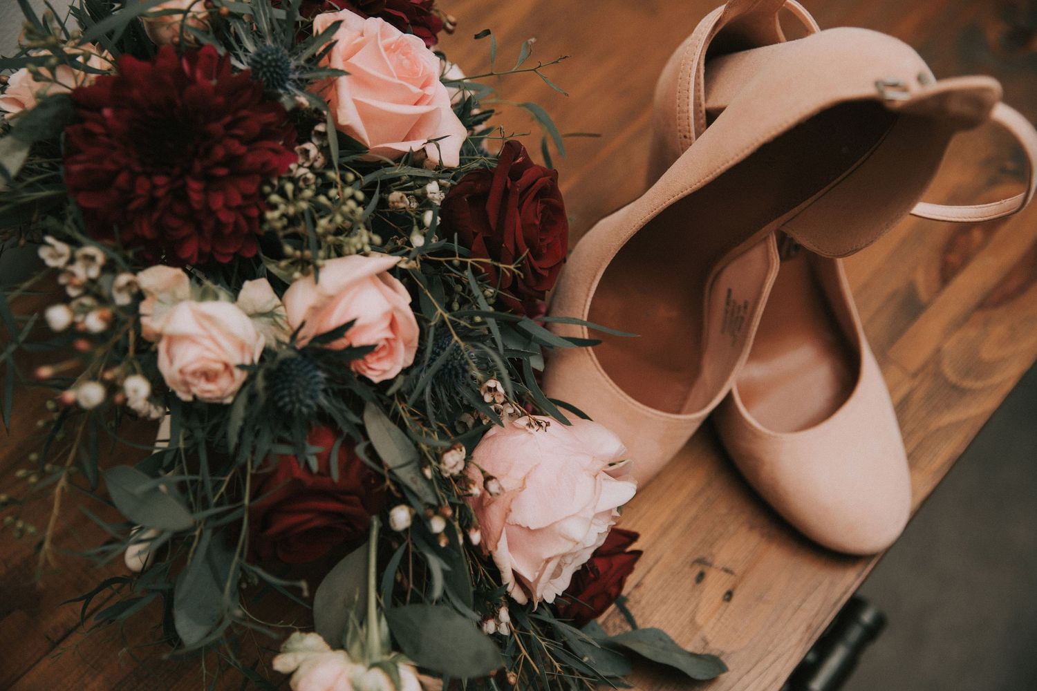 Bridal bouquet with pink and burgundy roses beside nude ballet flat wedding shoes on a wooden surface.