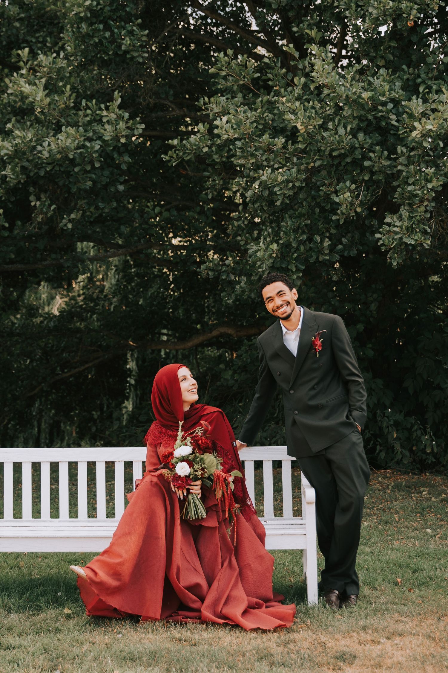 Muslim bride in red hijab and gown sits on white bench with groom in dark suit holding bouquet outdoors.