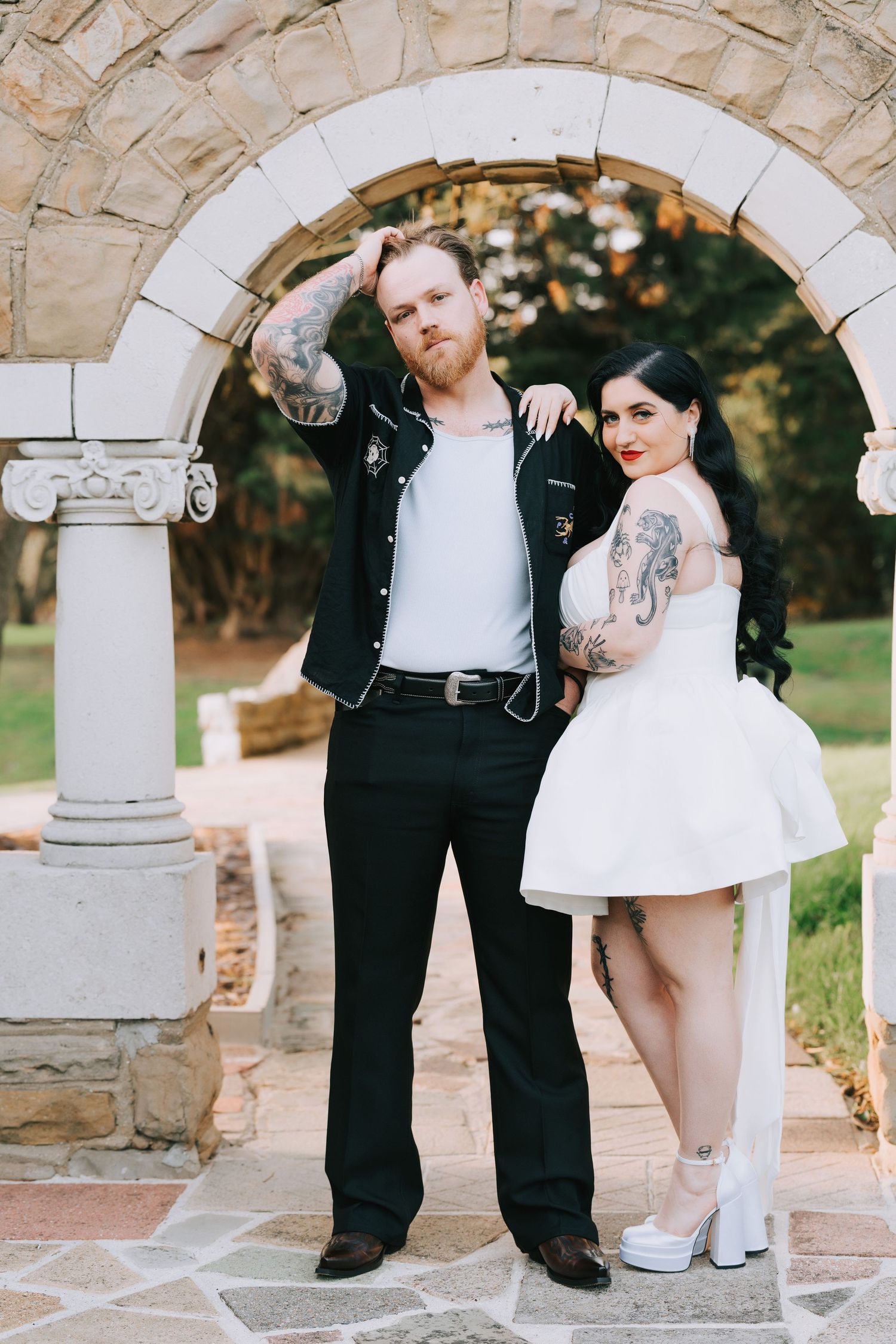 Tattooed couple in rockabilly wedding attire posing under stone arch, bride in white mini dress, groom in black jacket.