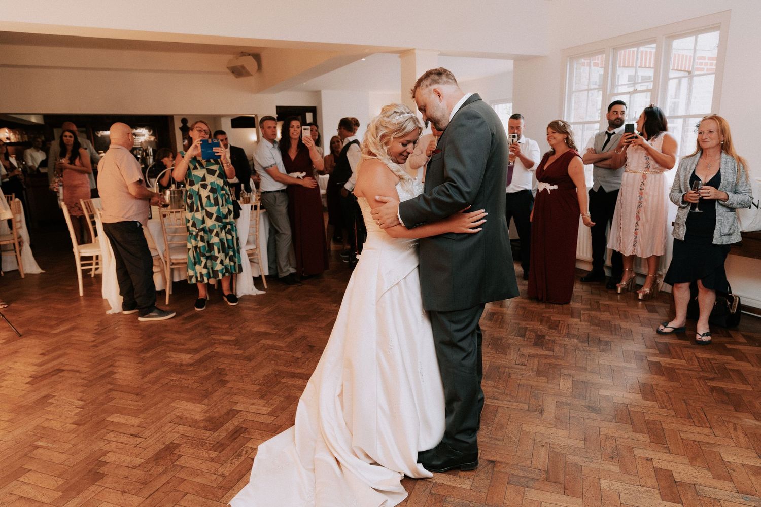 Bride and groom share their first dance at wedding reception on herringbone wood floor surrounded by guests.