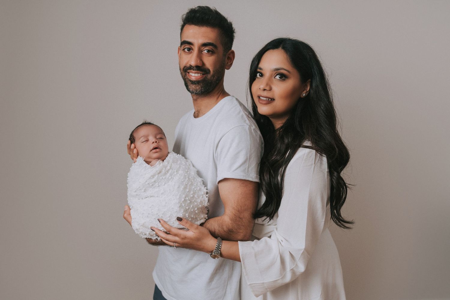 A family wearing white outfits poses together against a neutral grey background in a minimalist studio setting.